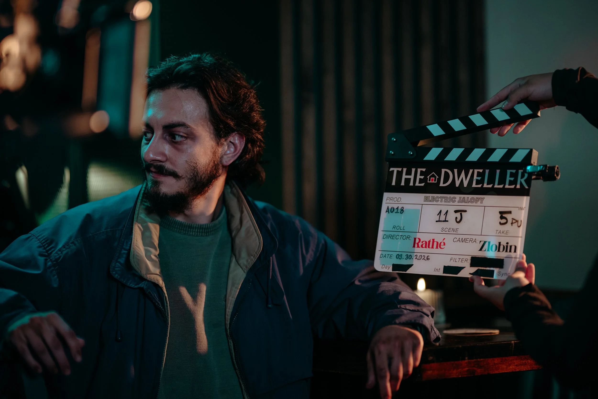 A man with dark, curly hair and a beard holds a film slate with details written on it. Part of a person is visible on the right, reaching towards the film slate. The background is dark with some bokeh lights.