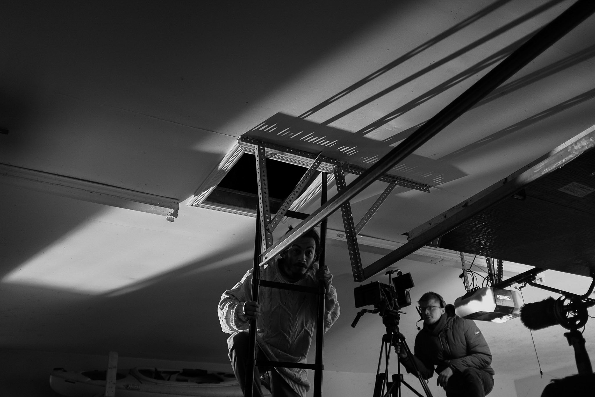 People working on film or photography equipment indoors, with part of a ceiling and an open ceiling hatch visible, illuminated by a lamp.