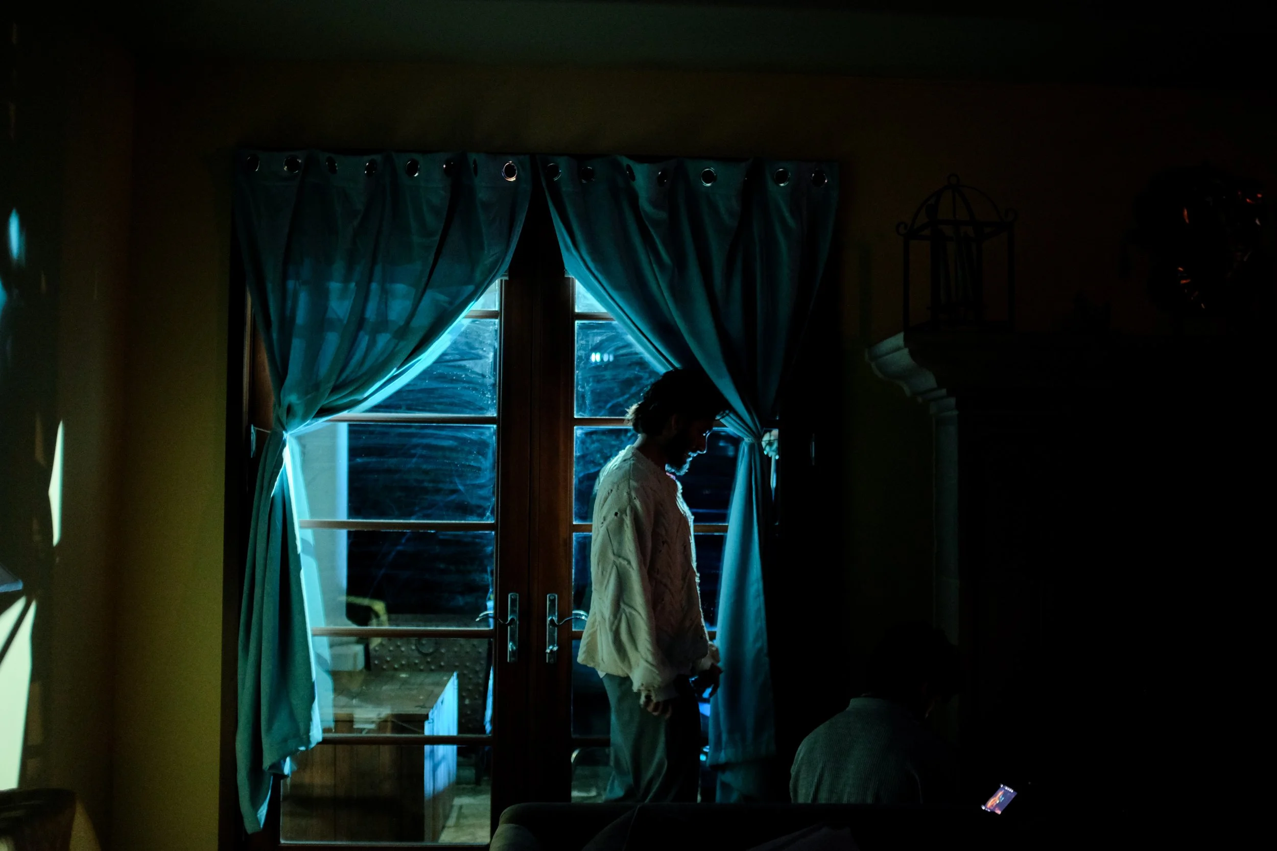 A man with a beard standing near a window with blue curtains, inside a dimly lit room.
