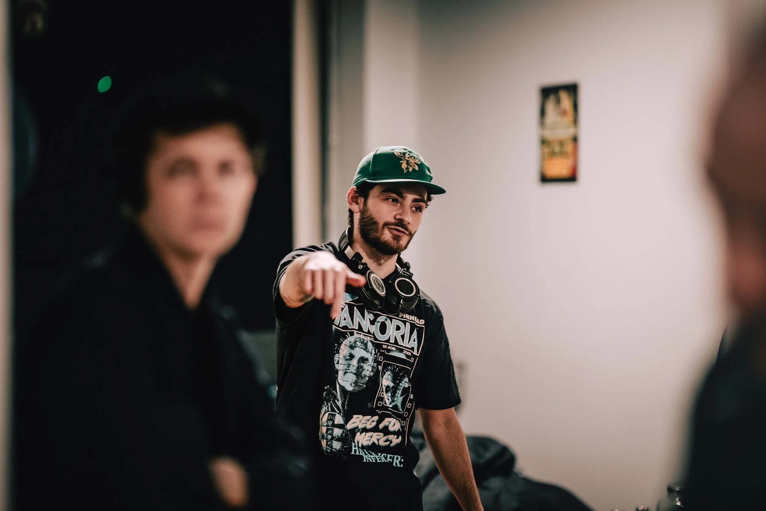 A young man with a beard and dark hair wearing a green baseball cap with a deer logo, black headphones around his neck, and a graphic T-shirt, is pointing towards the camera, engaged in a conversation or activity in an indoor setting.