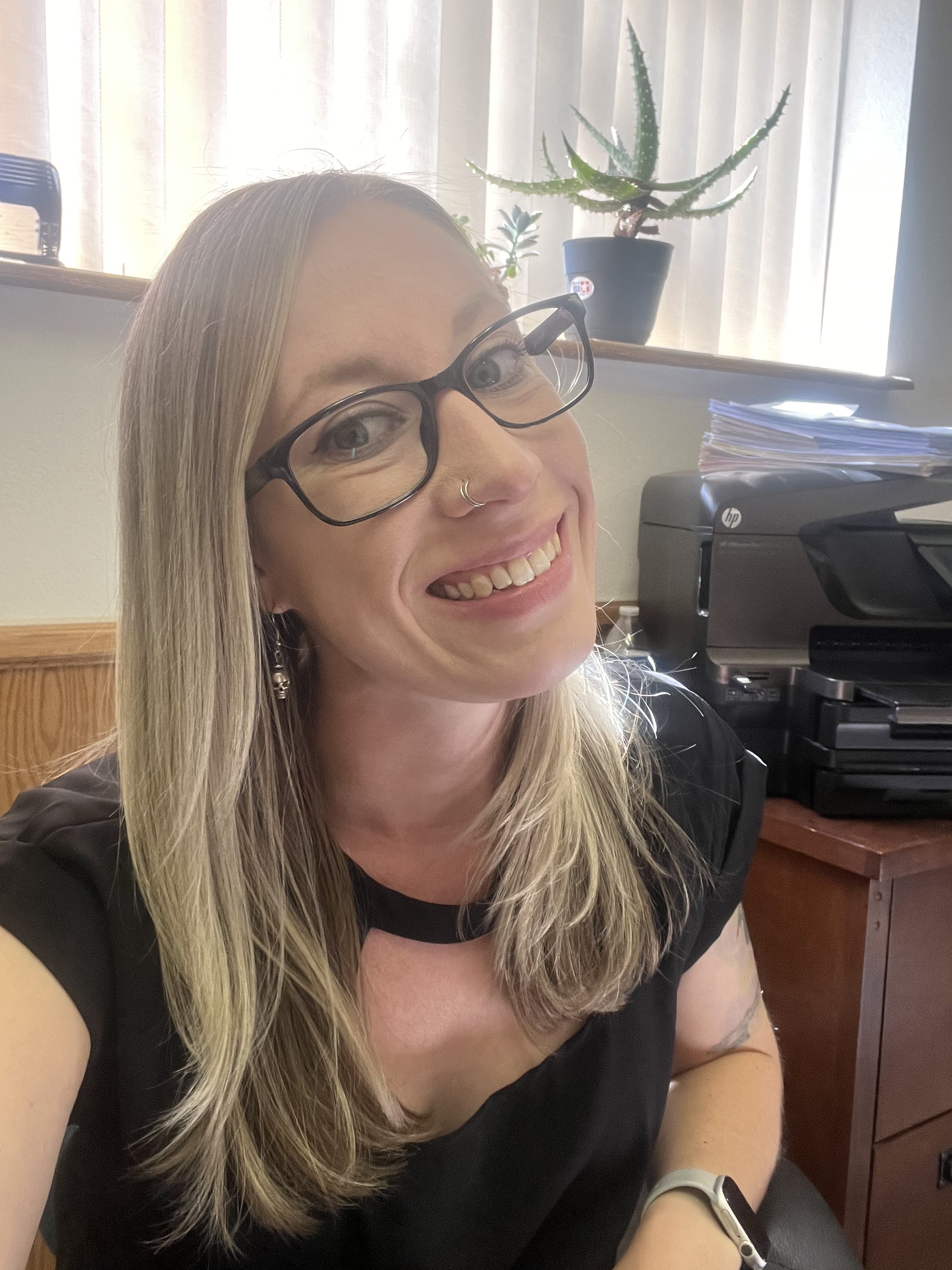 A smiling woman with long blonde hair, wearing black glasses and a nose ring, sitting in an office with a printer, stack of papers, and aloe vera plant in the background.