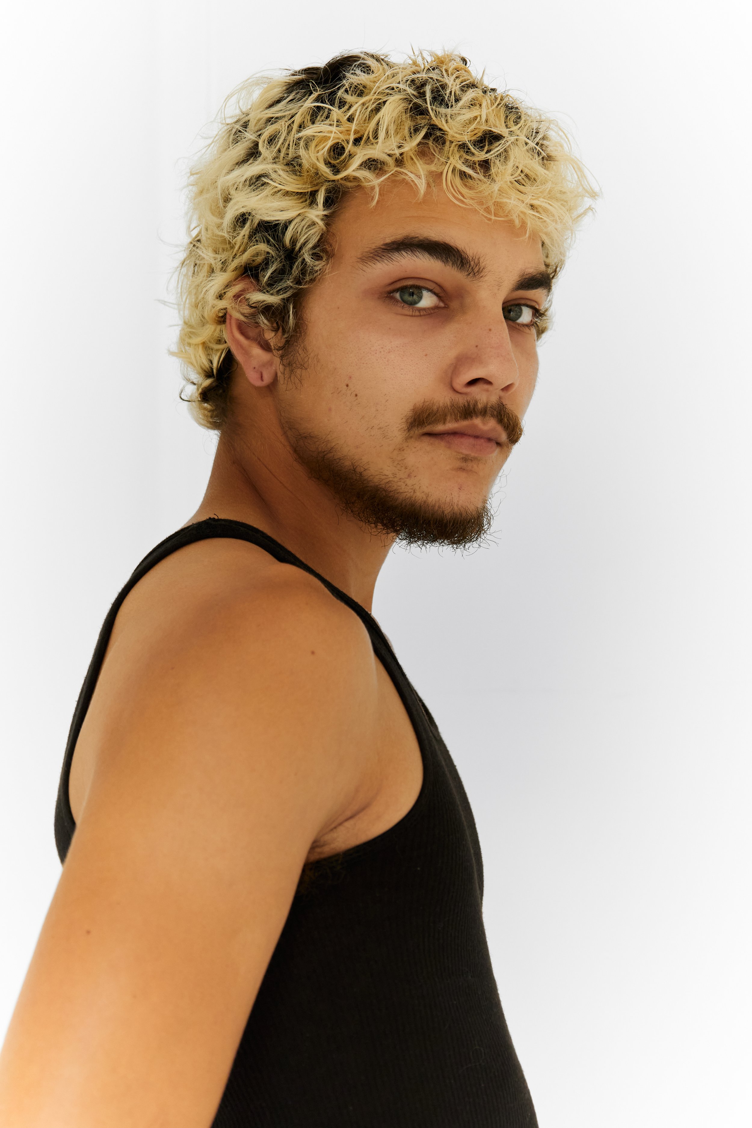 Quinton, male fashion model represented by Noirvelle Models, side portrait in a black tank top against a white studio background.
