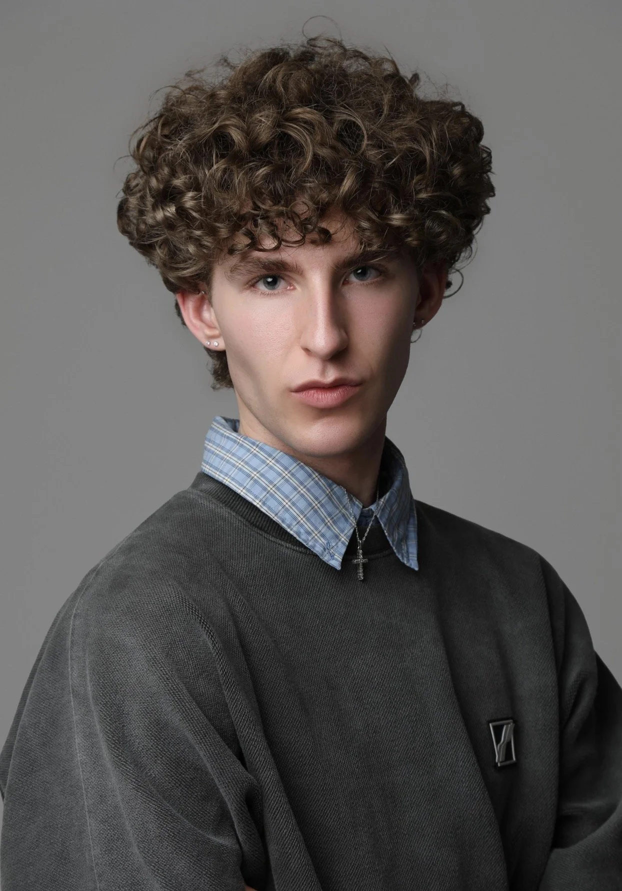 Young man with curly brown hair, blue eyes, wearing a dark gray sweatshirt over a light blue plaid shirt, silver earrings, and a cross necklace, posing against a plain gray background.