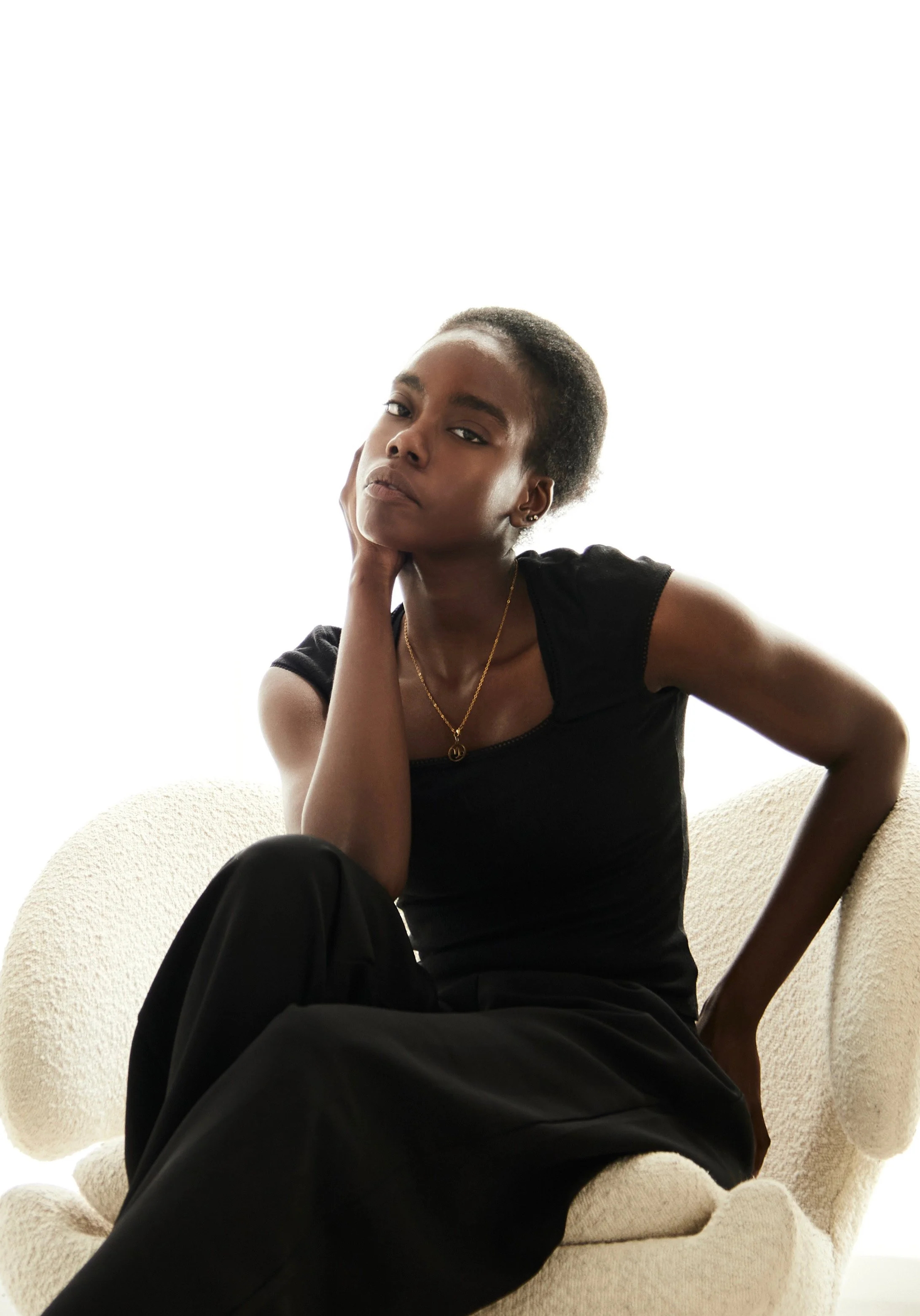  Daisy, female fashion model represented by Noirvelle Models, seated in a cream armchair wearing an all-black look.
