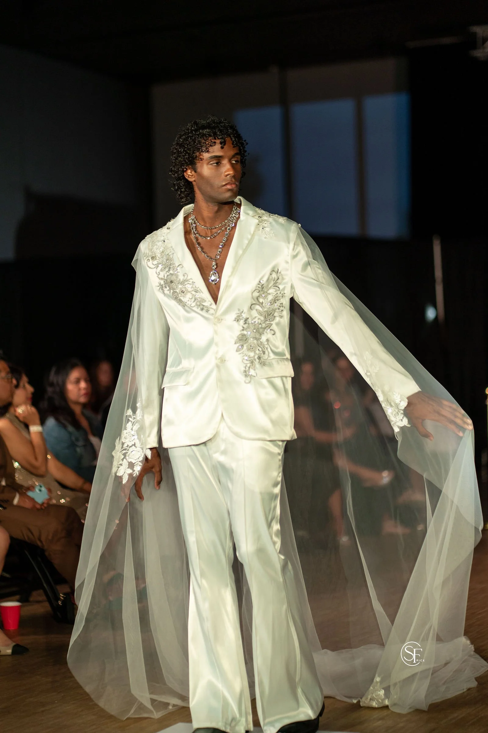 Xander, male fashion model represented by Noirvelle Models, walking the runway in a white embroidered suit during a fashion show.
