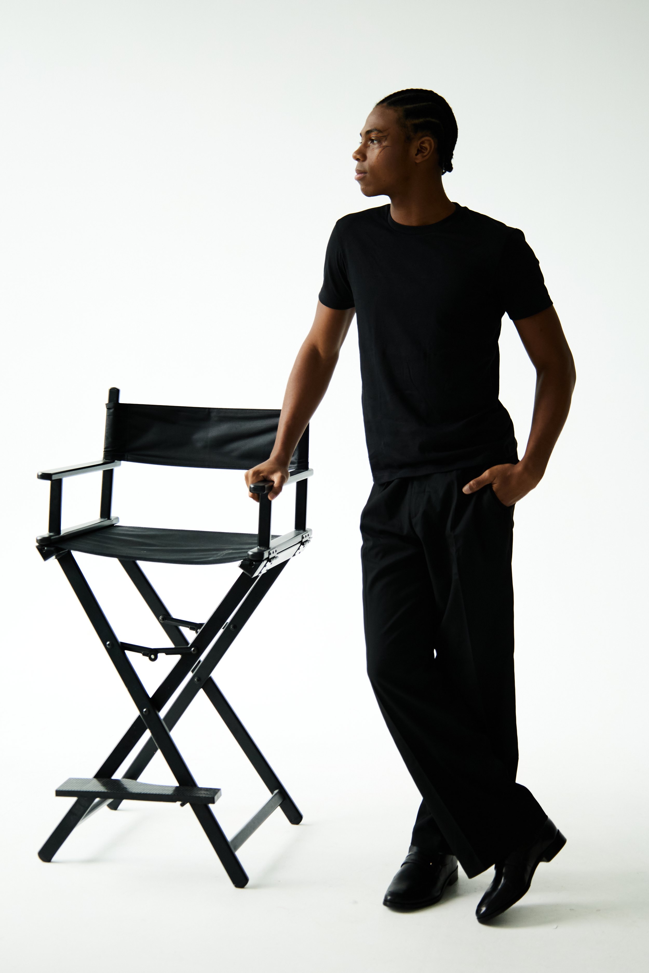 Jahvante, male fashion model represented by Noirvelle Models, standing beside a director’s chair in a black outfit during a studio photoshoot.
