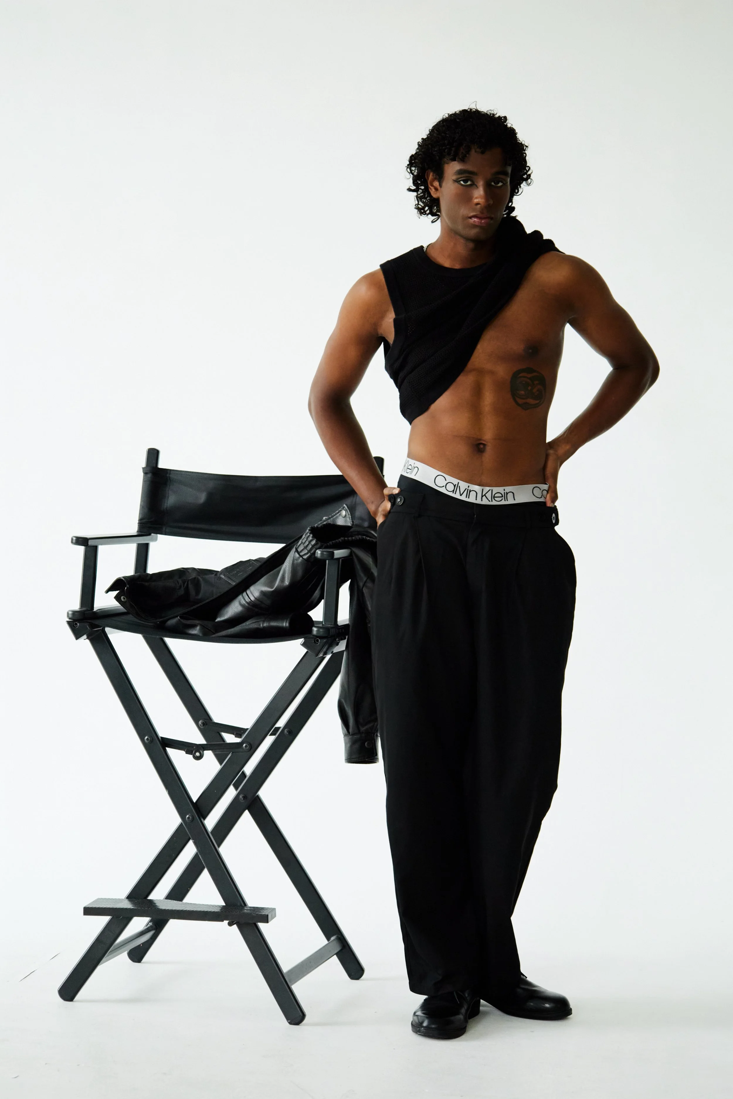  Xander, male fashion model represented by Noirvelle Models, posing in a black fashion look beside a director’s chair.