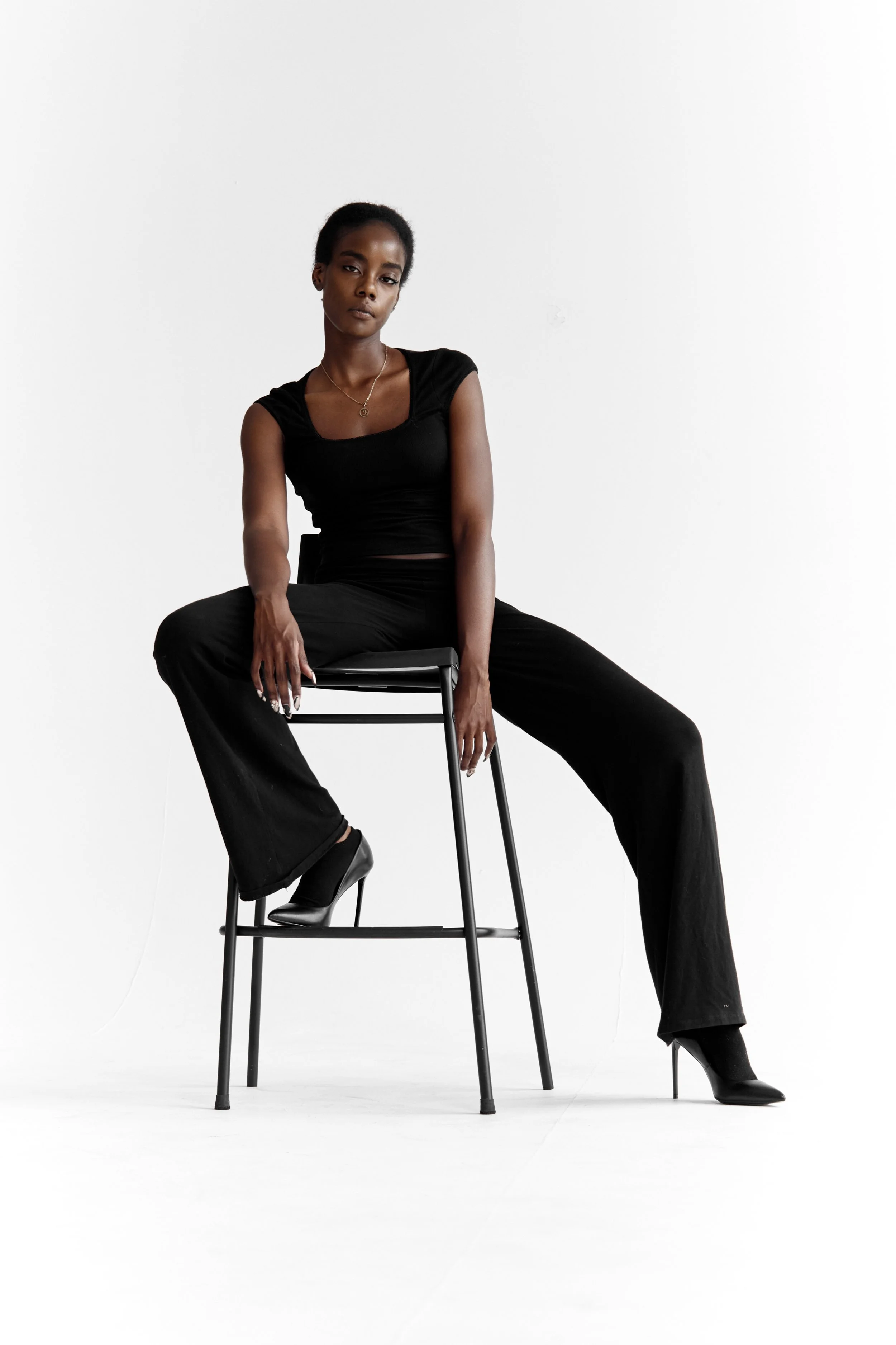  Daisy, female fashion model represented by Noirvelle Models, seated on a high chair in an all-black look against a white studio background.

