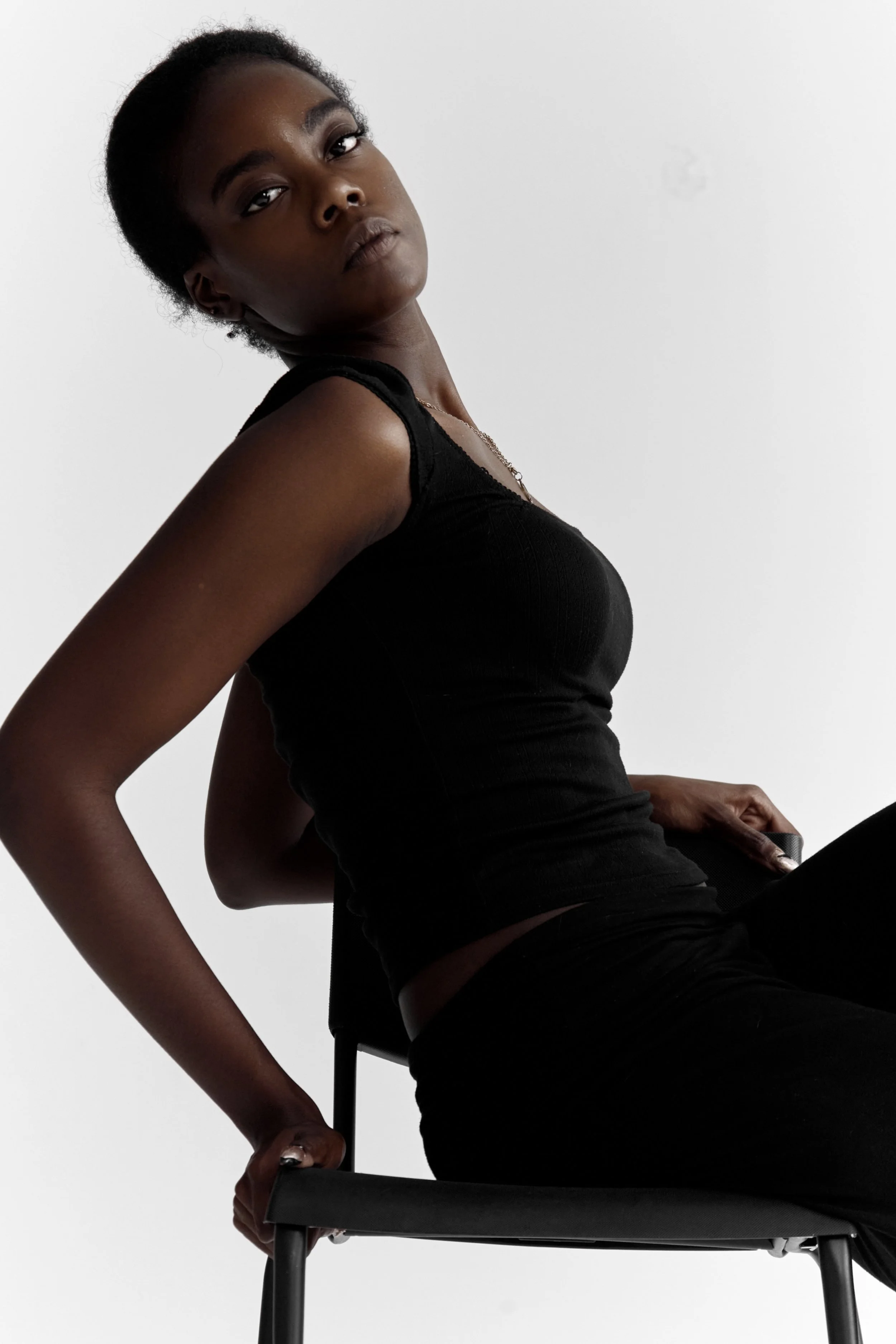 Daisy, female fashion model represented by Noirvelle Models, posing sideways on a black chair against a white studio background.