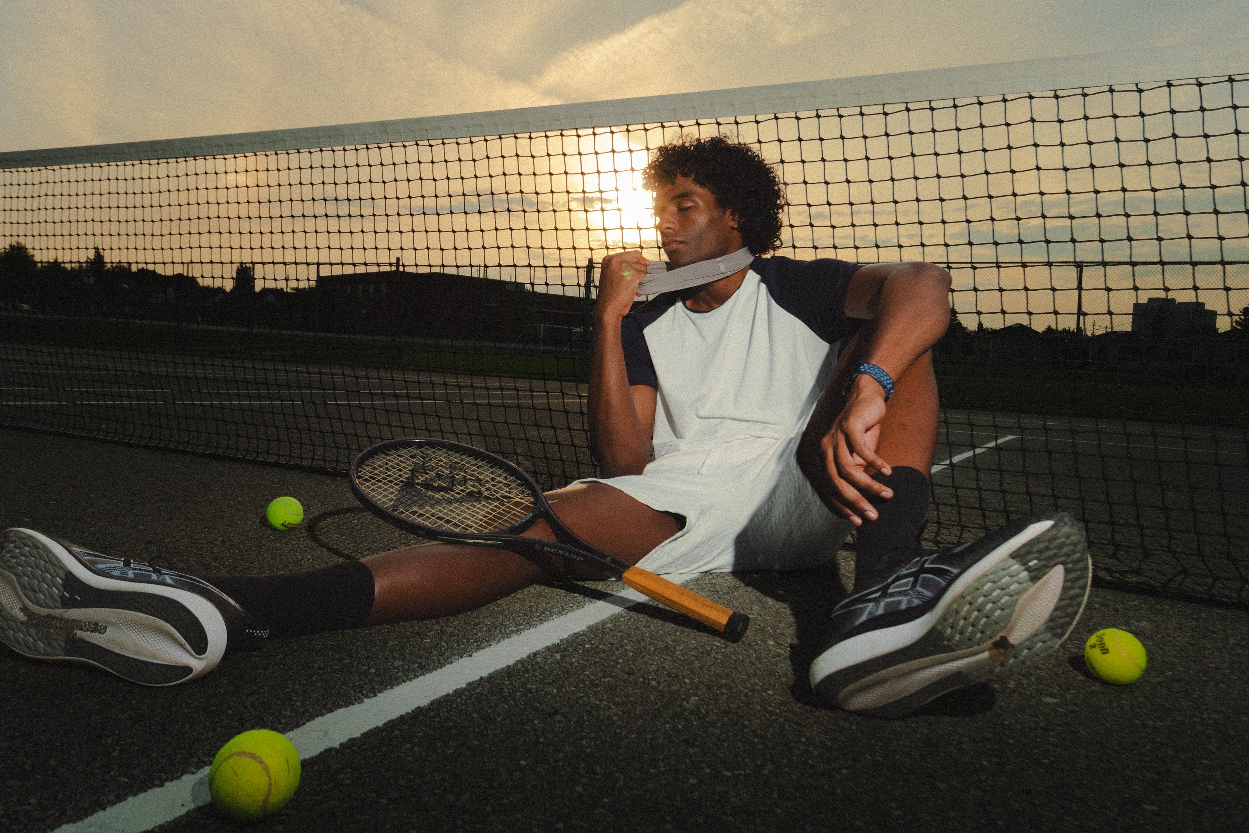 Xander, male fashion model represented by Noirvelle Models, posing with a tennis racket on a tennis court at sunset.
