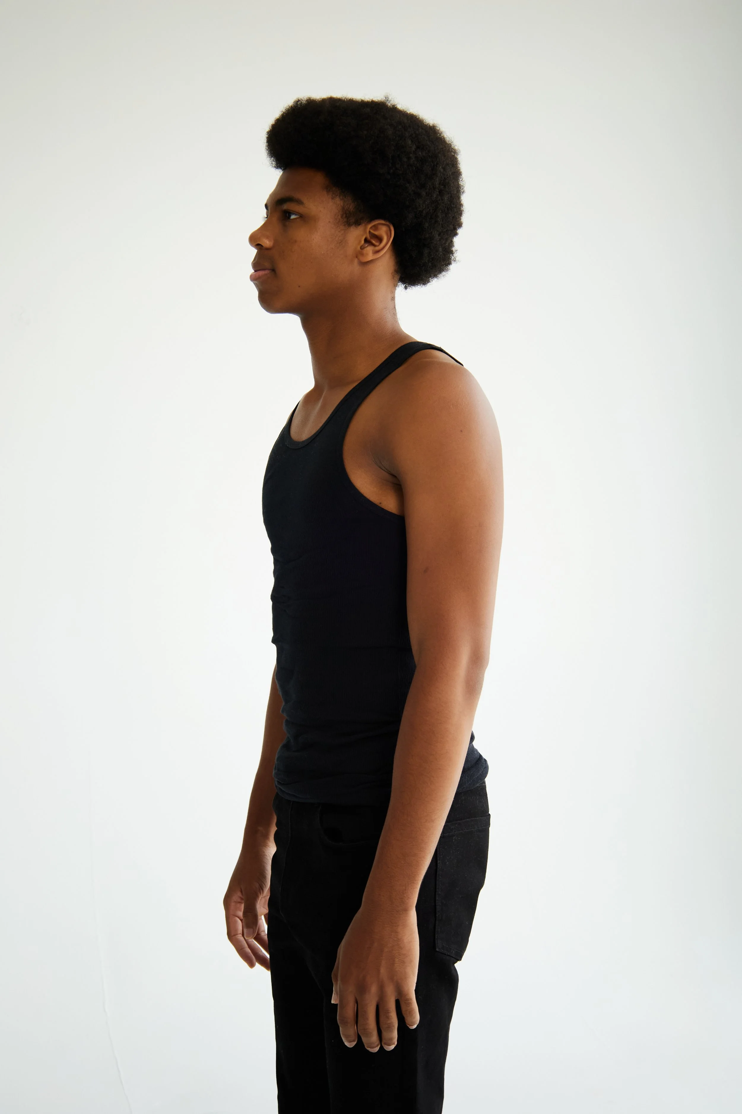 Jahvante, male fashion model represented by Noirvelle Models, side profile in a black sleeveless top and black pants against a white studio background.