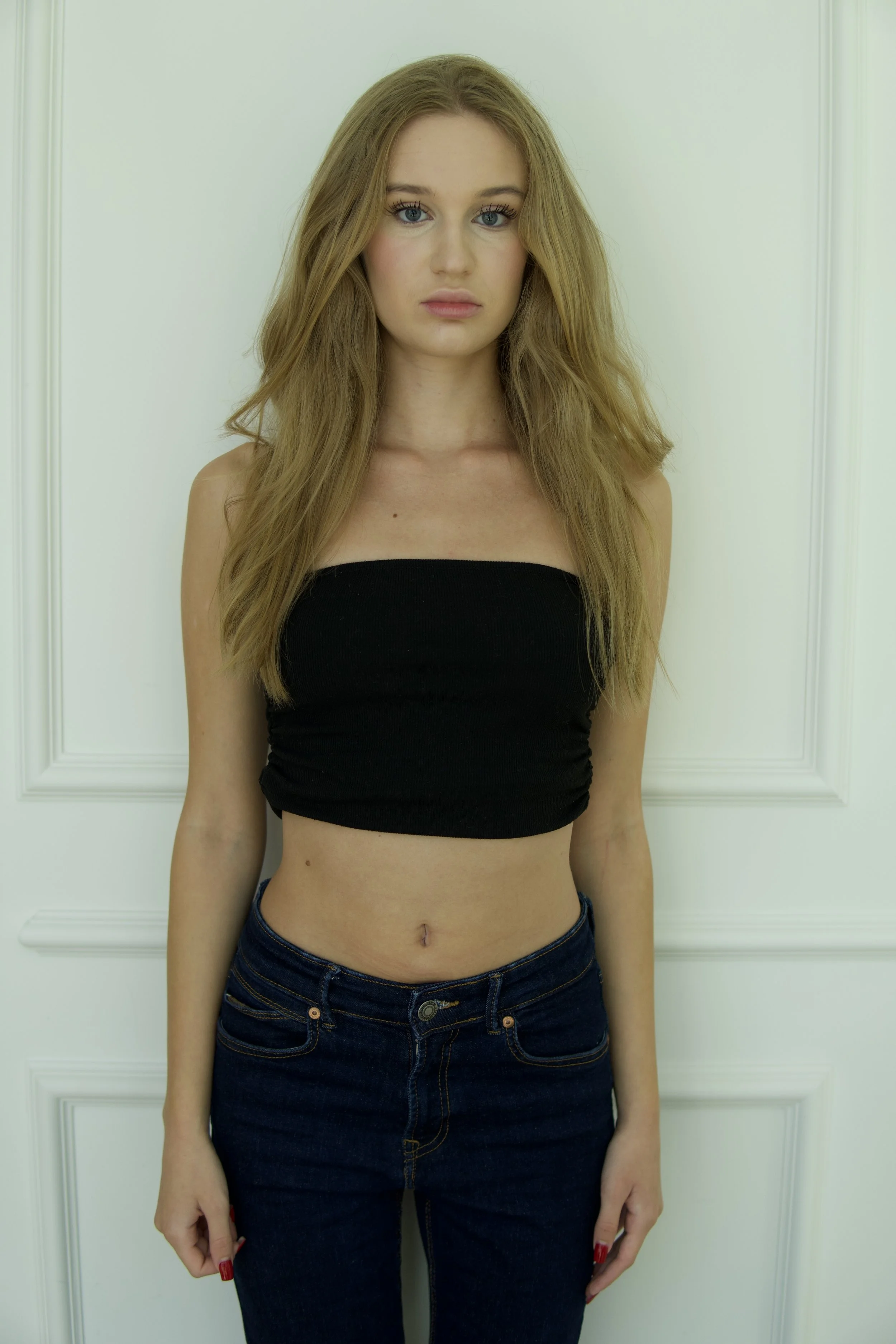 Victoria, female fashion model represented by Noirvelle Models, wearing a black crop top and dark jeans against a white paneled wall.
