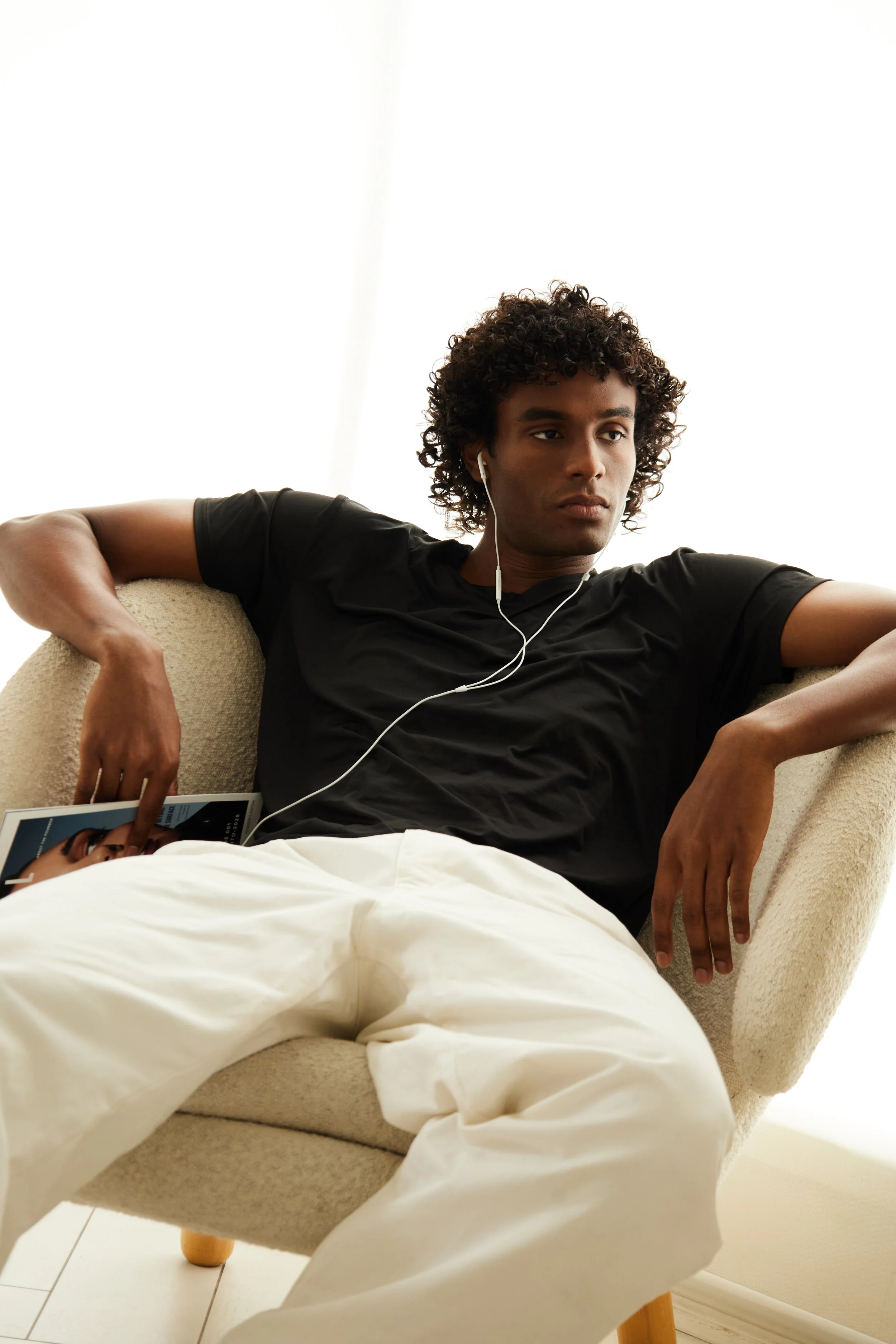Xander, male fashion model represented by Noirvelle Models, seated in a beige armchair wearing a black shirt and white pants.
