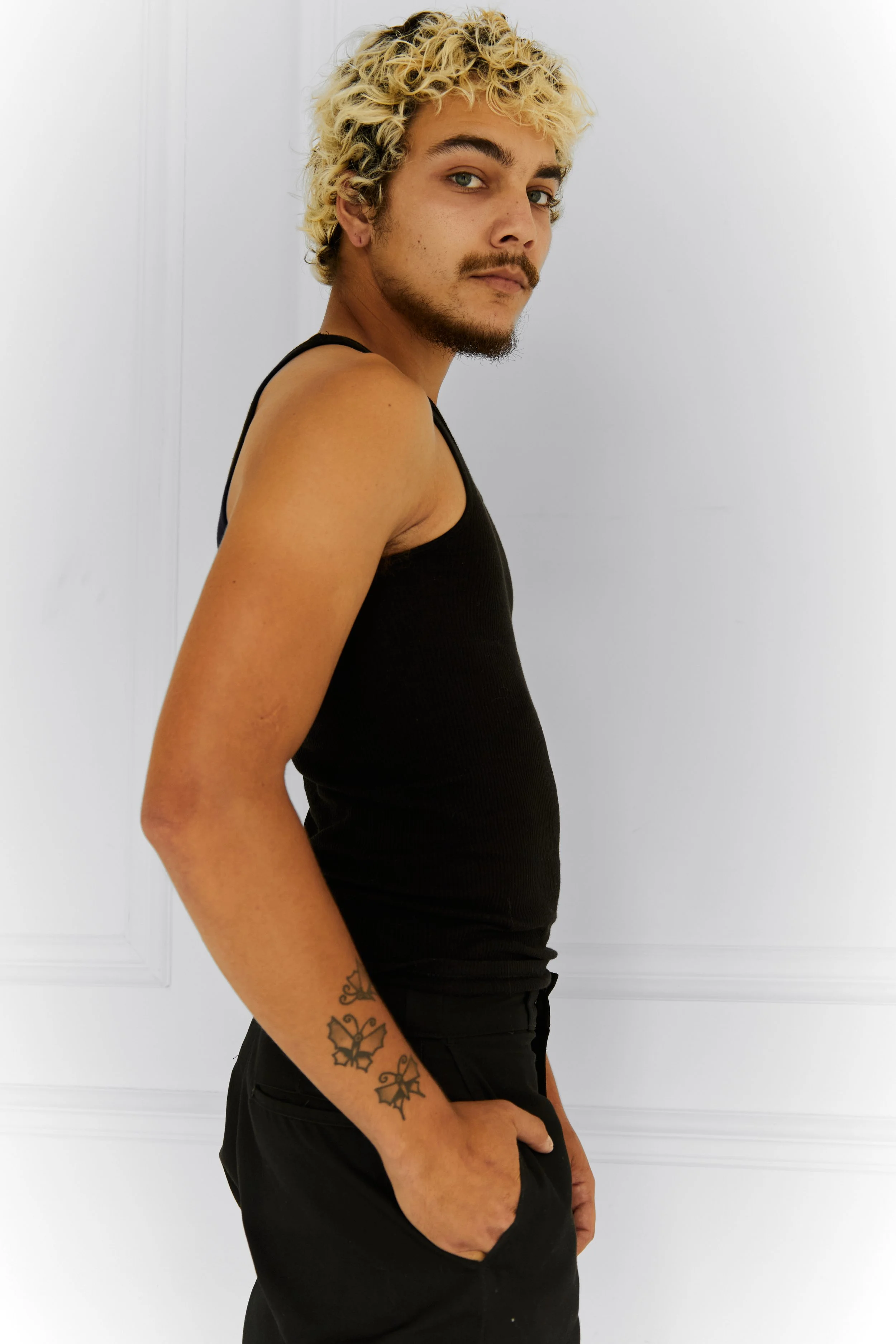Quinton, male fashion model represented by Noirvelle Models, posing in a black tank top and black pants against a white studio background.