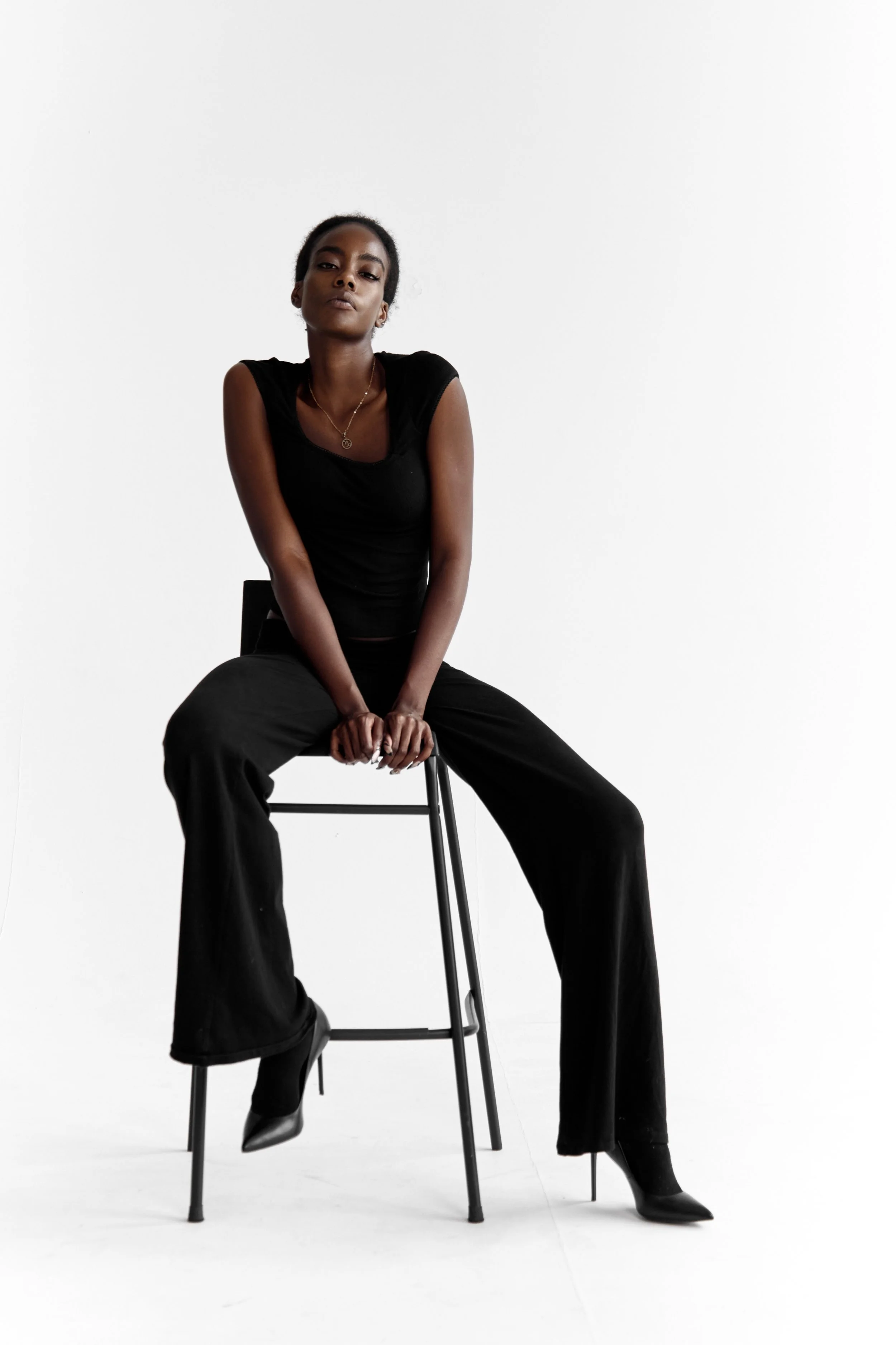A woman sitting on a black stool with a white background, wearing black pants, a black sleeveless top, and black high-heeled boots. She has short hair, a necklace, and looks confidently at the camera.