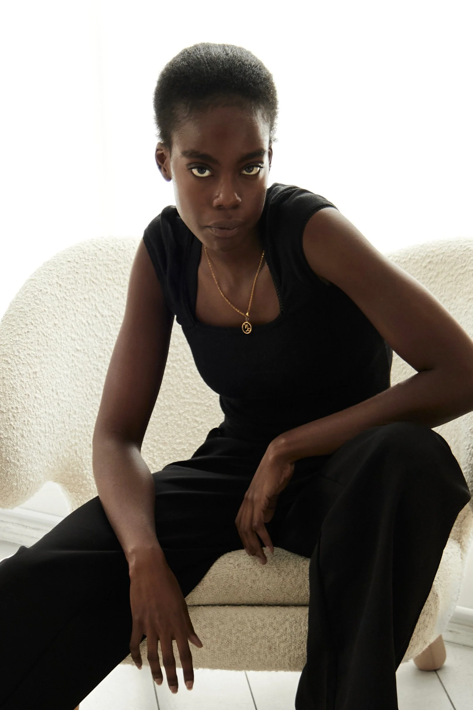  Daisy, female fashion model represented by Noirvelle Models, seated on a light sofa wearing a black outfit and gold necklace.

