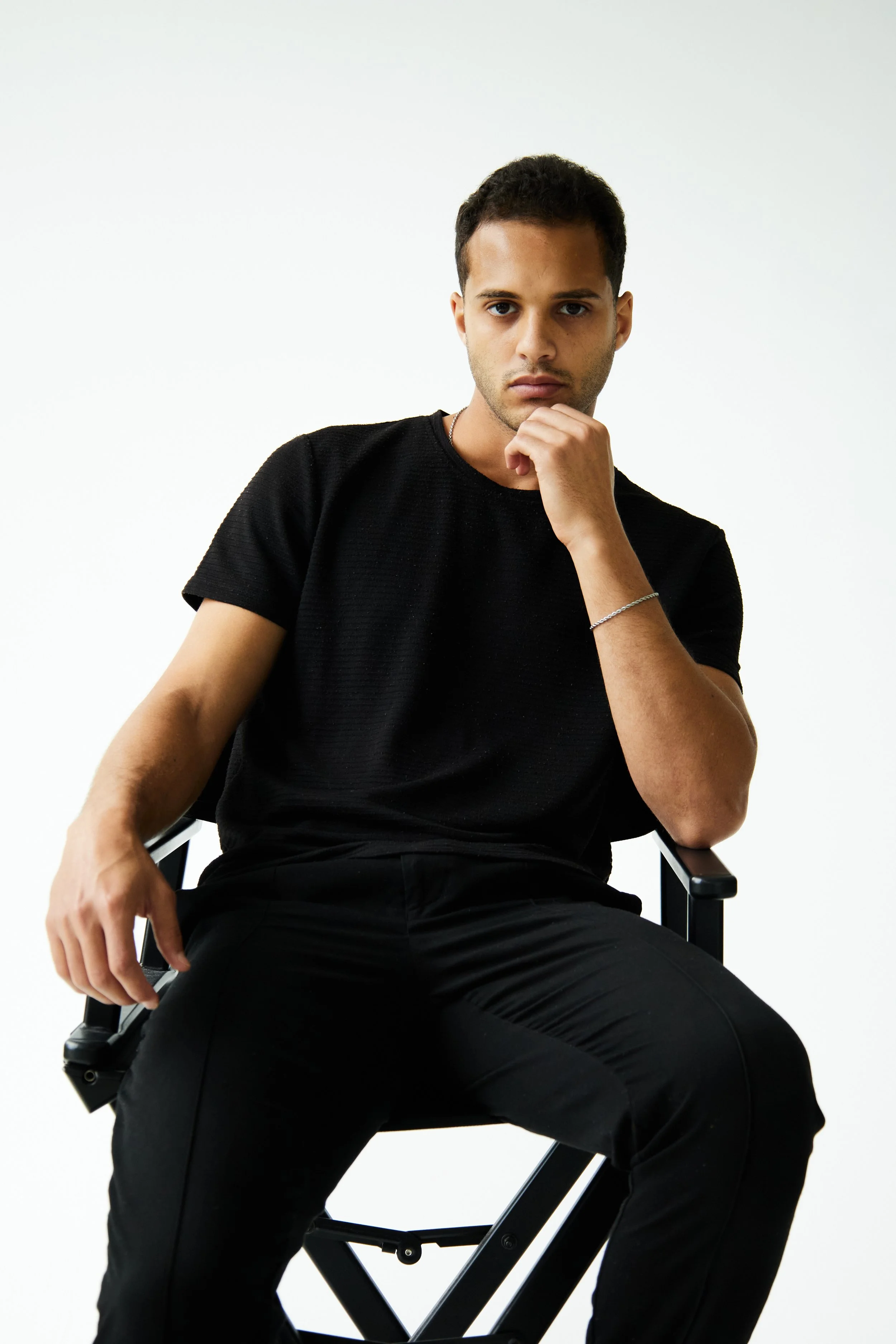 Bashir, male fashion model represented by Noirvelle Models, seated on a director’s chair during a minimalist studio photoshoot.
