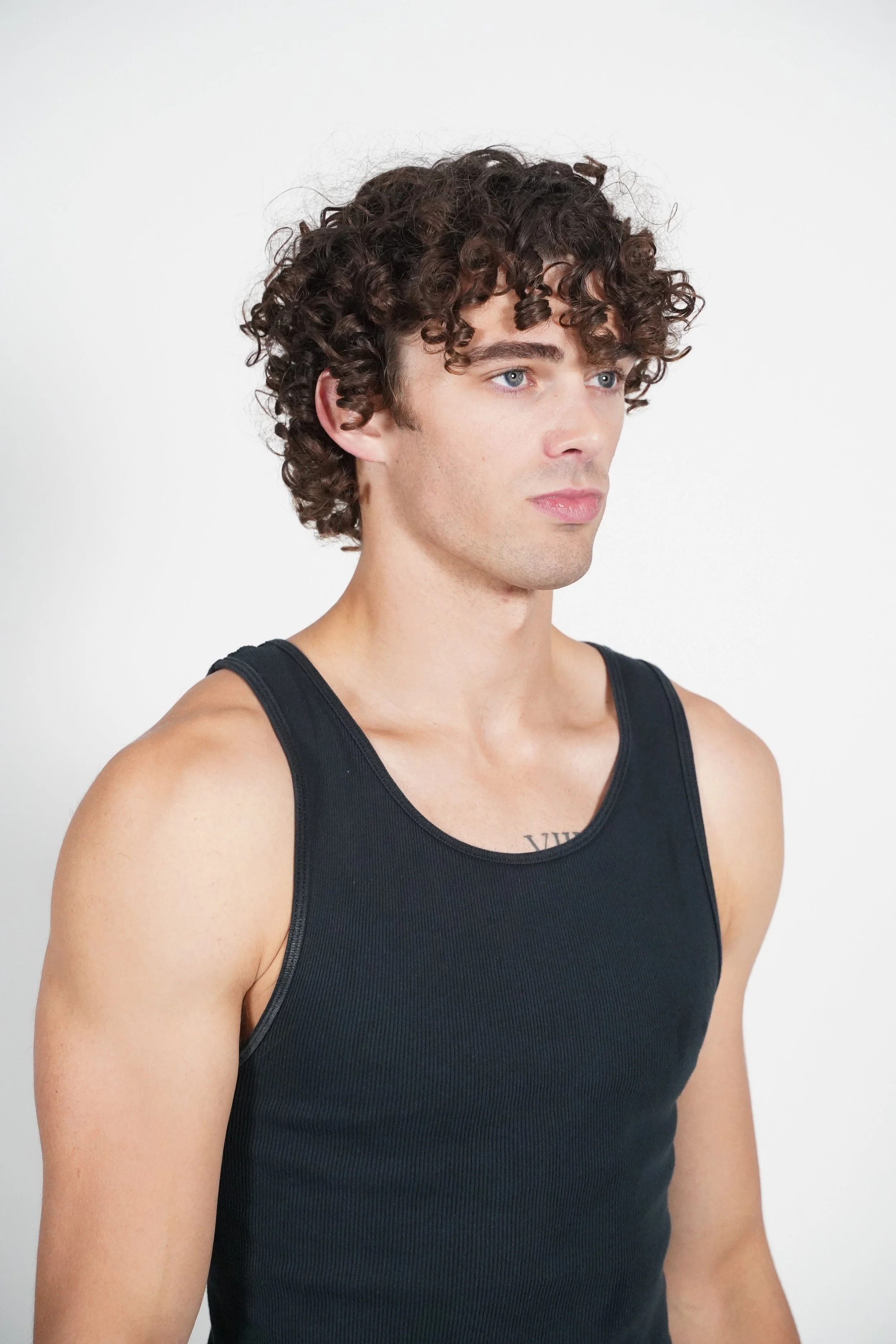  Maverick, male fashion model represented by Noirvelle Models, posing in profile in a black sleeveless top against a white studio background.