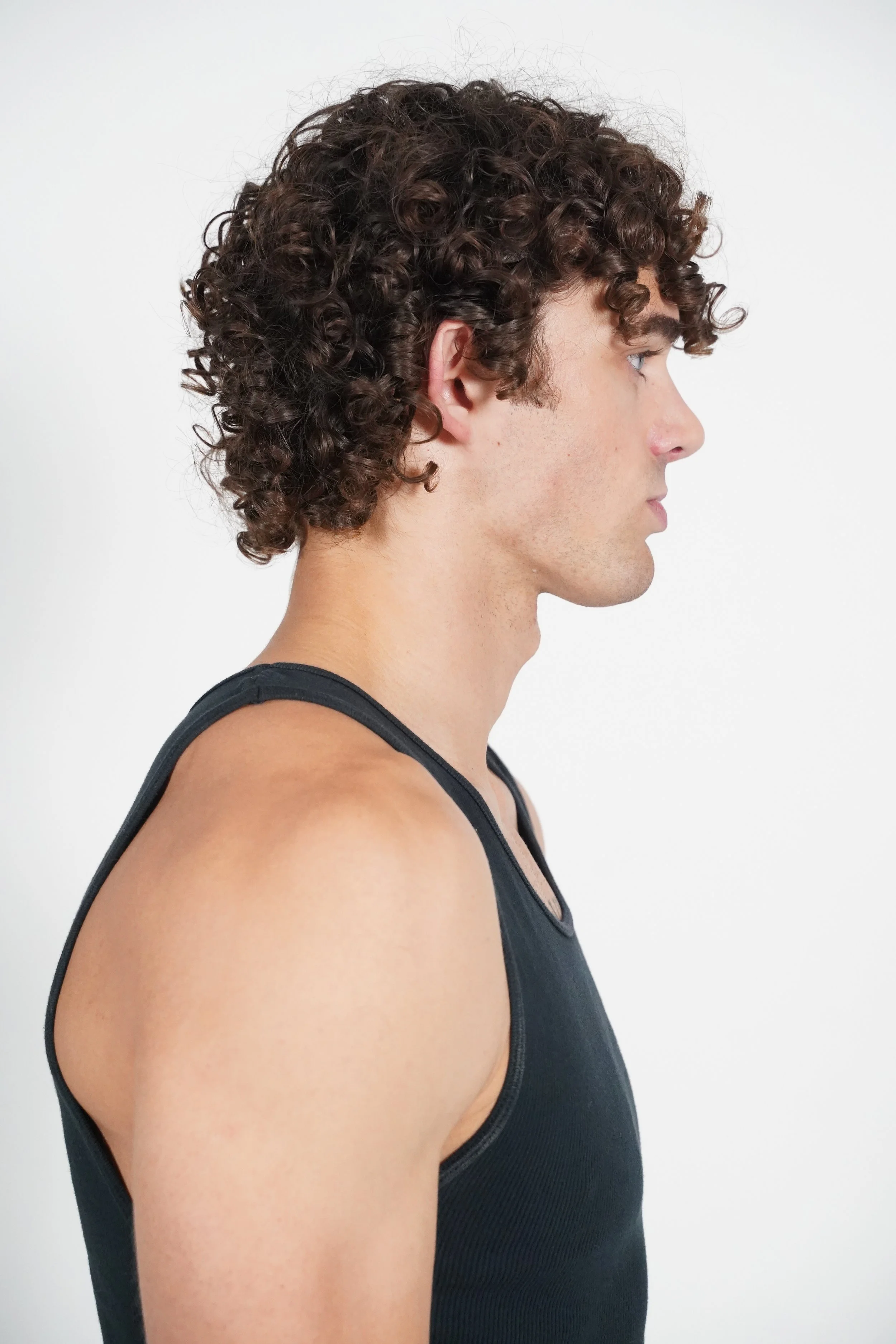 Maverick, male fashion model represented by Noirvelle Models, posing in profile in a black sleeveless top against a white studio background.
