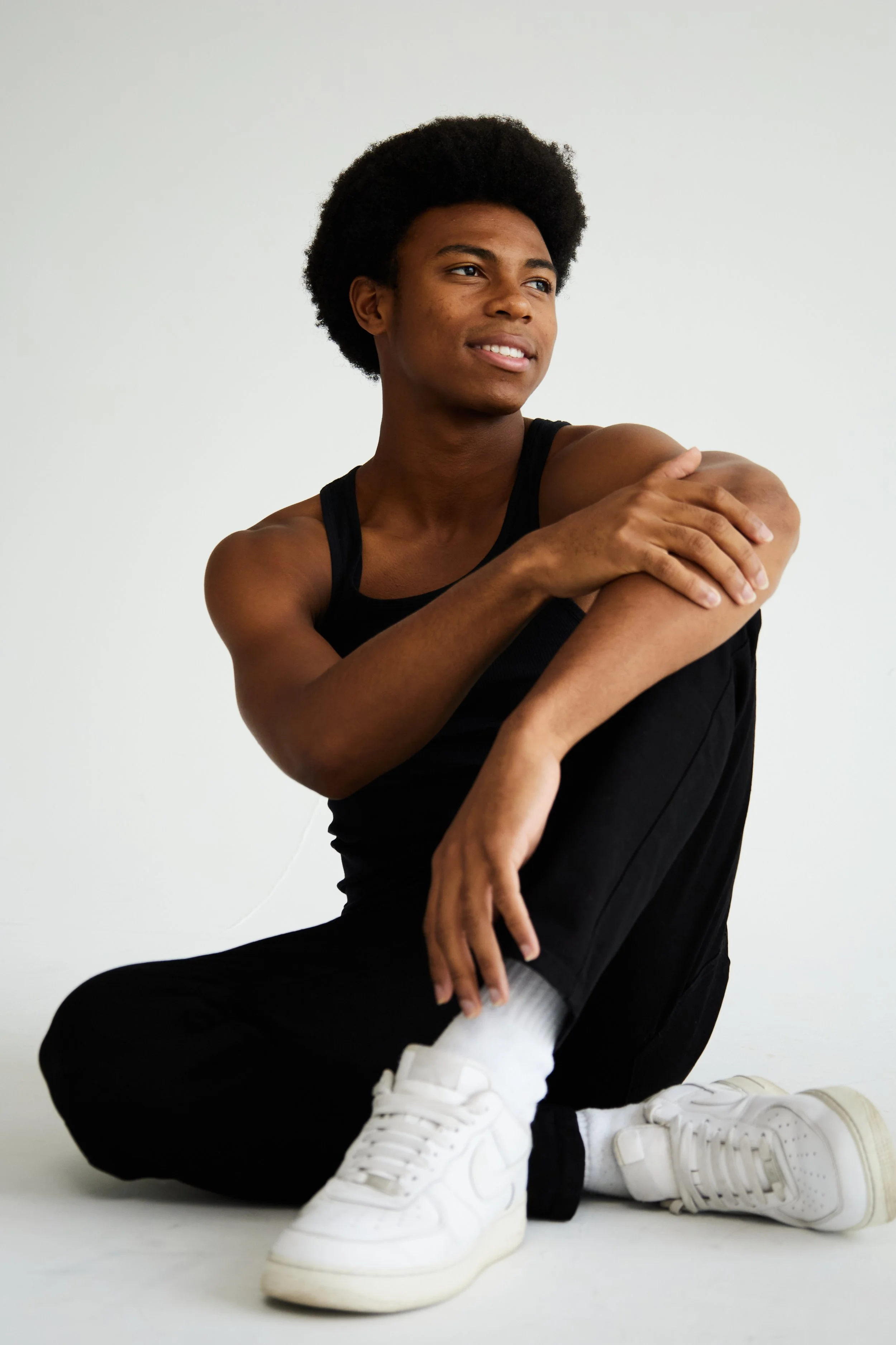 Jahvante, male fashion model represented by Noirvelle Models, seated on the floor in a black outfit against a white studio background.

