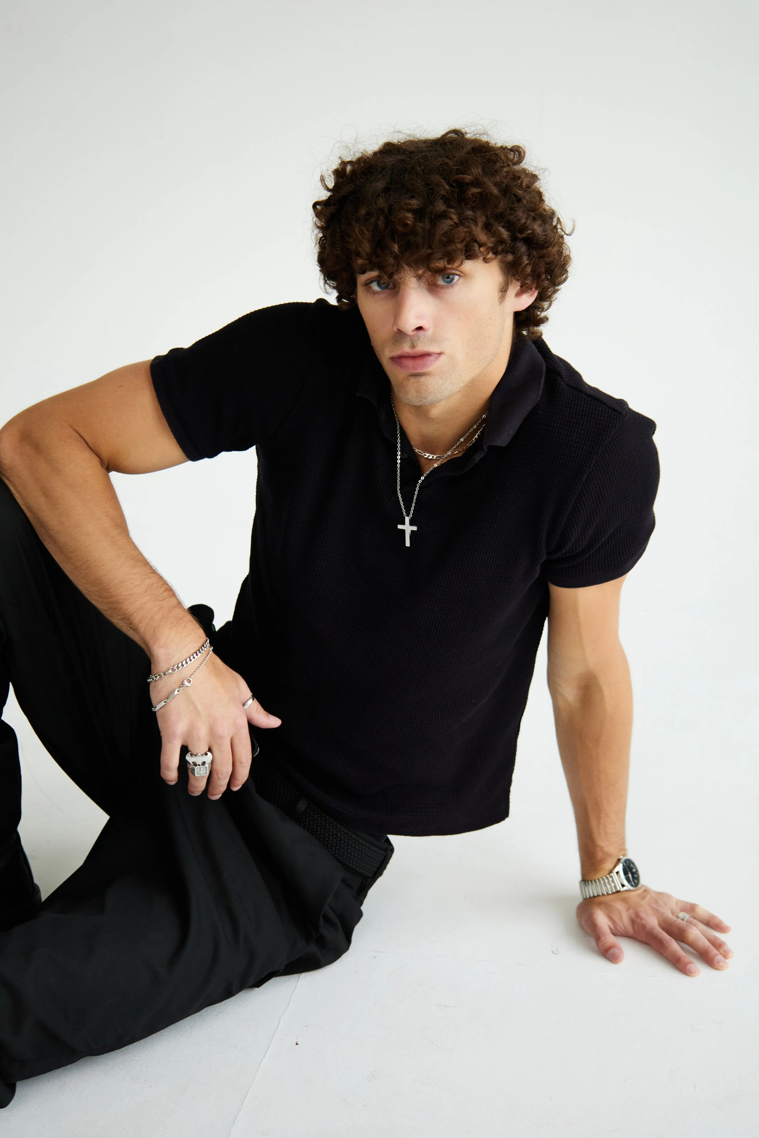 Maverick, male fashion model represented by Noirvelle Models, seated on the floor in an all-black look against a white studio background.