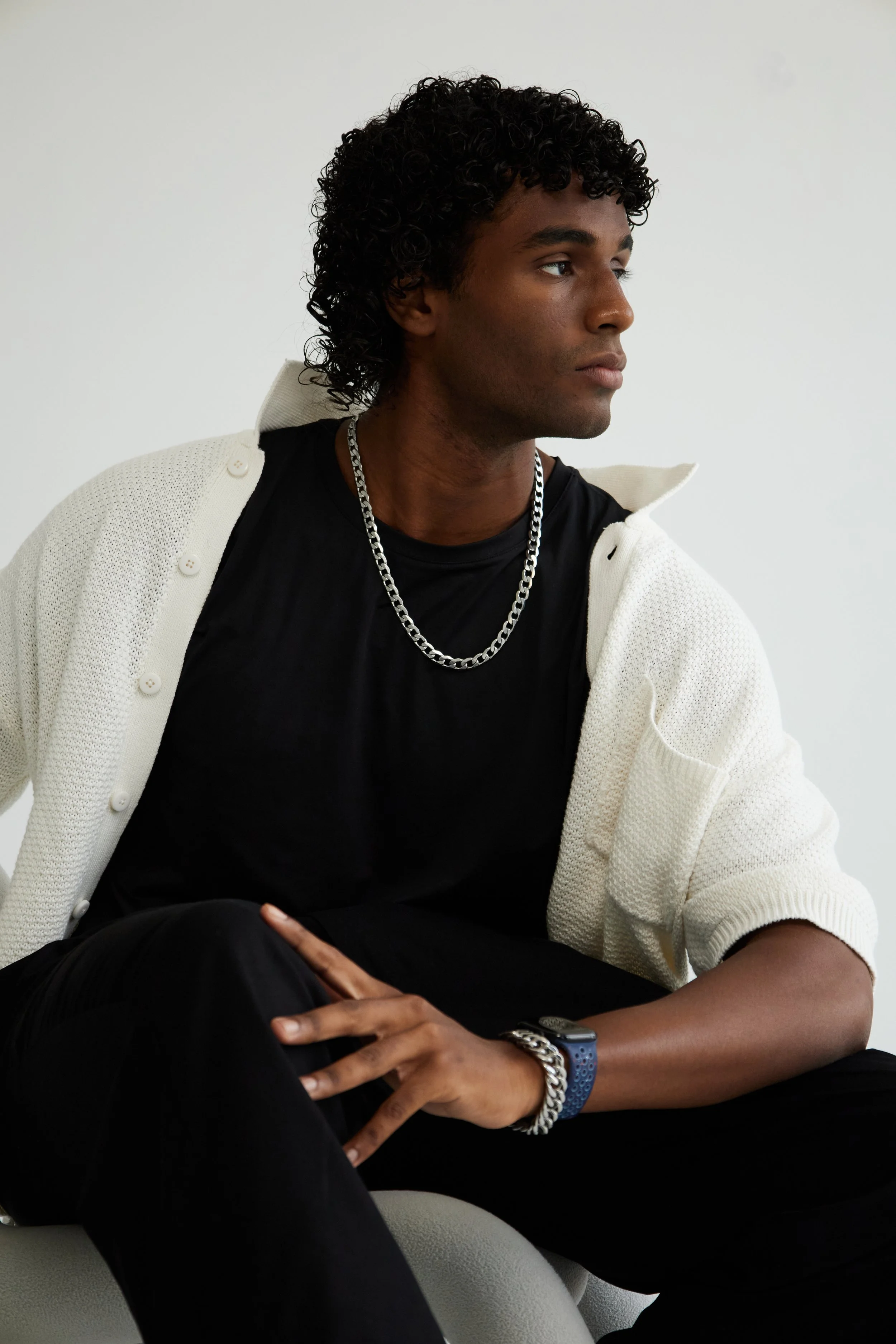 Xander, male fashion model represented by Noirvelle Models, seated in a black T-shirt and white cardigan against a white studio background.