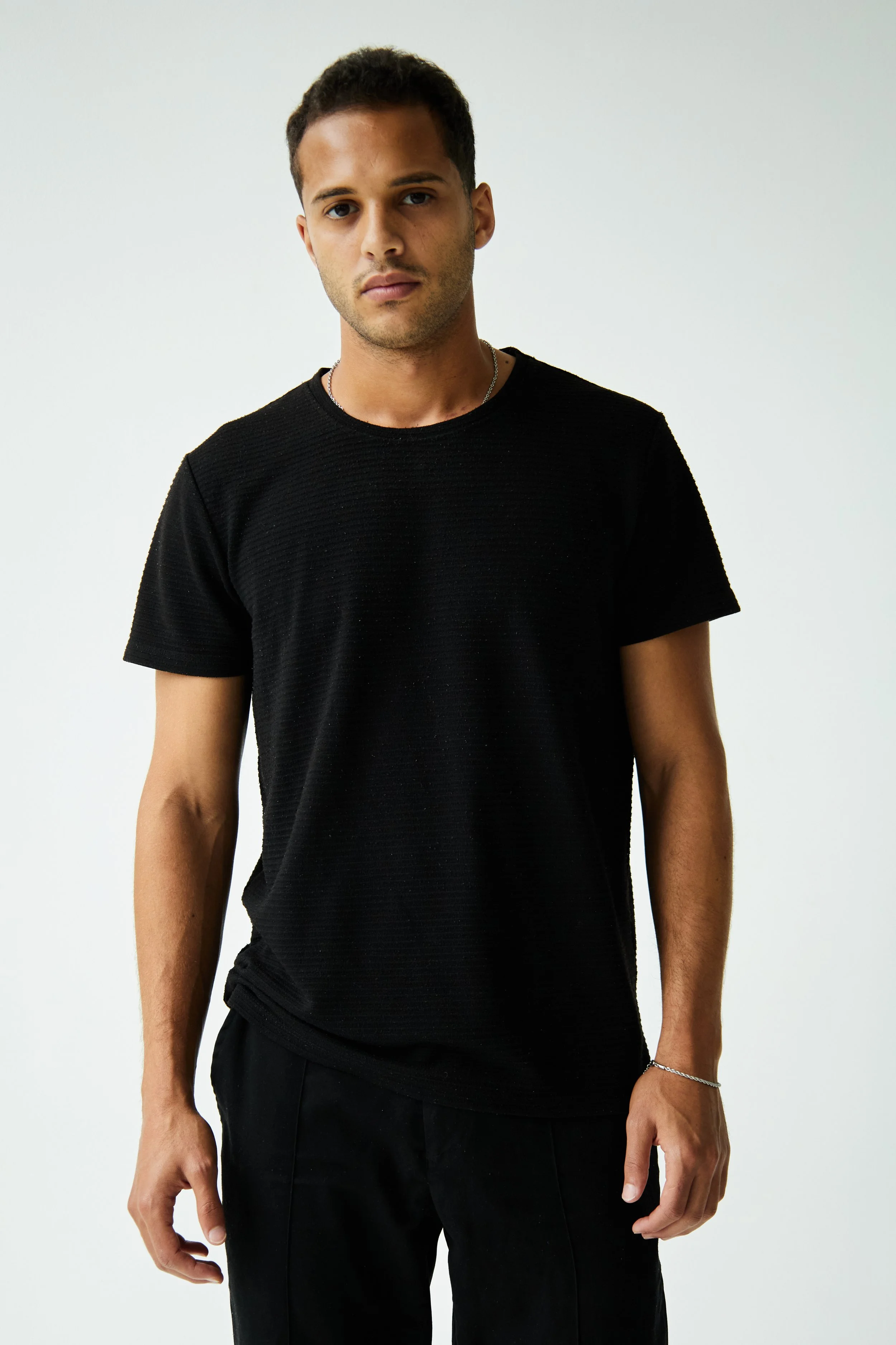  Bashir, male model represented by Noirvelle Models, standing in a black T-shirt and black pants against a white studio background.
