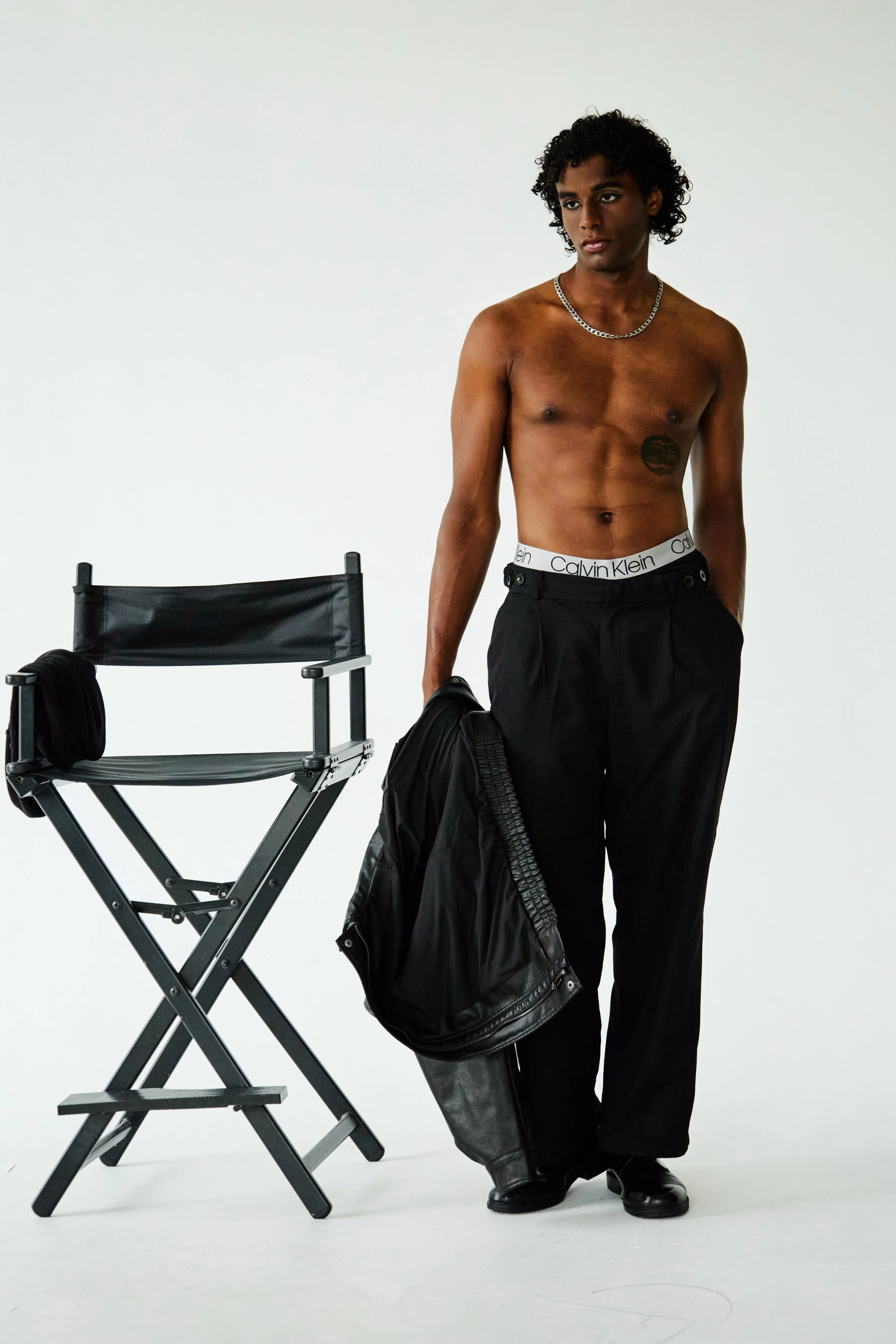 Xander, male fashion model represented by Noirvelle Models, posing shirtless in black jeans beside a director’s chair.
