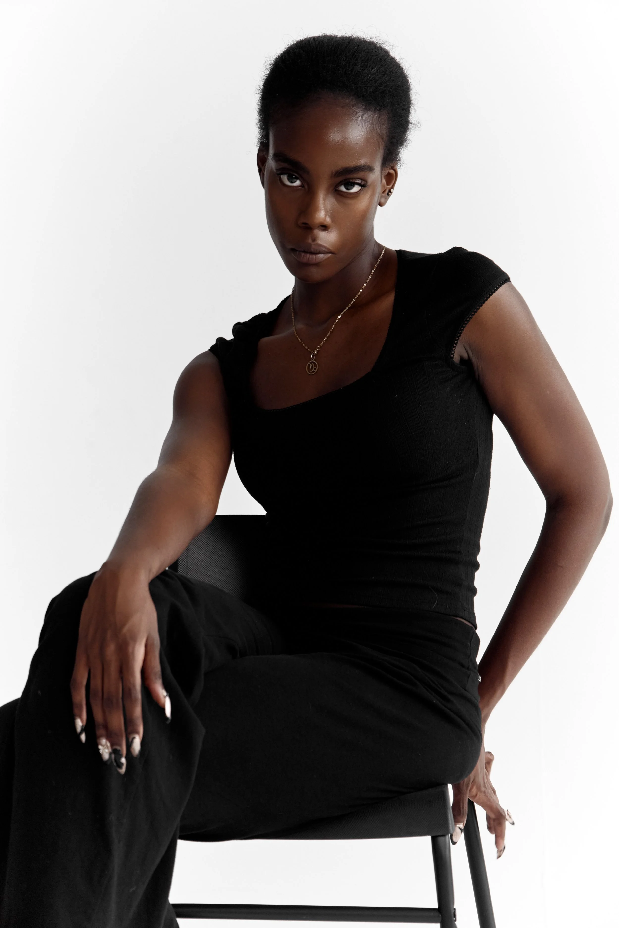  Daisy, female fashion model represented by Noirvelle Models, seated in a black outfit and jewelry against a white studio background.