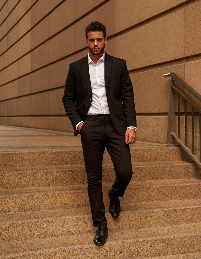 Bashir, male fashion model represented by Noirvelle Models, walking outdoors in a black suit and white shirt.