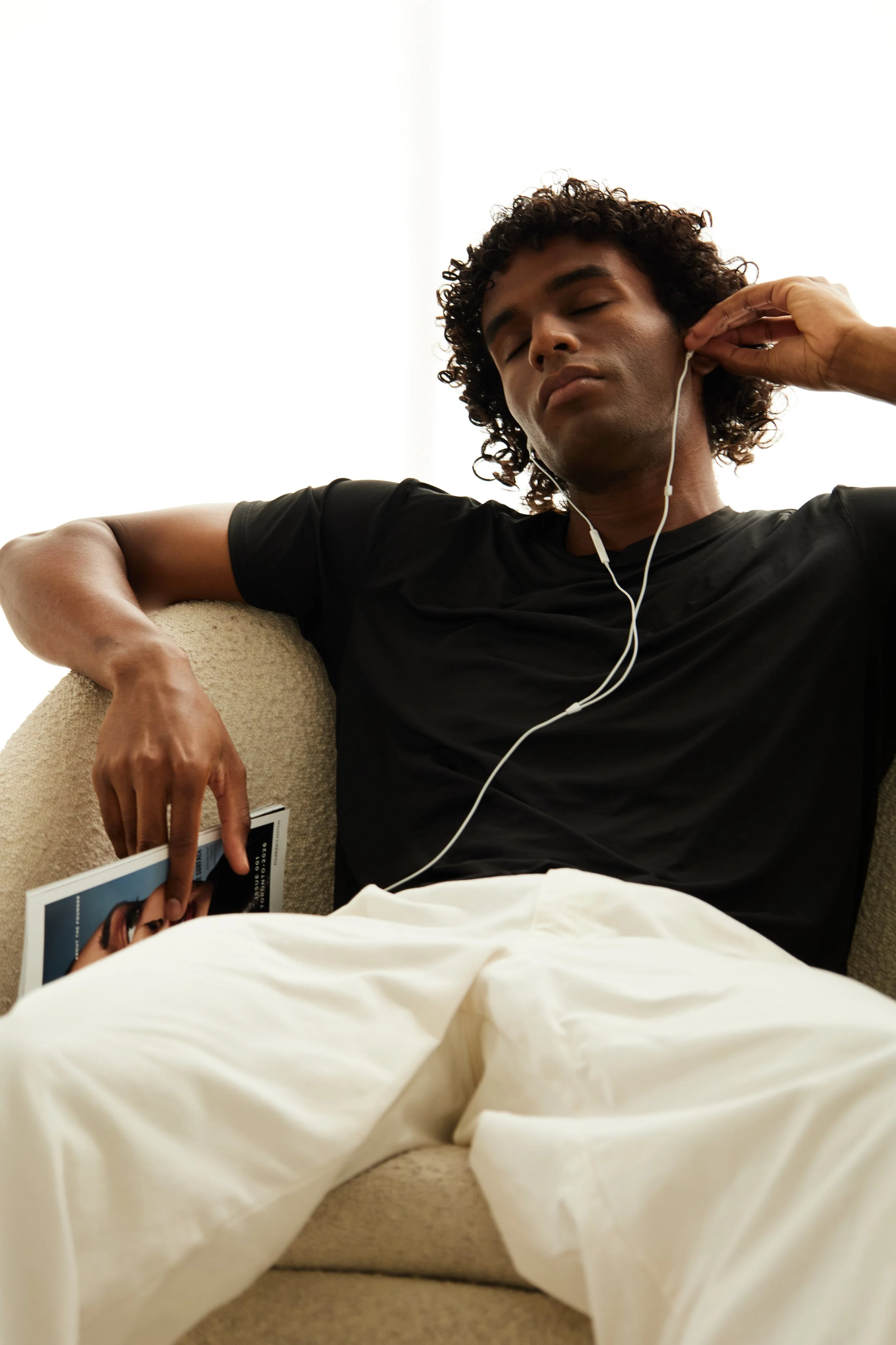 Xander, male fashion model represented by Noirvelle Models, relaxing on a beige sofa with a book and earphones.
