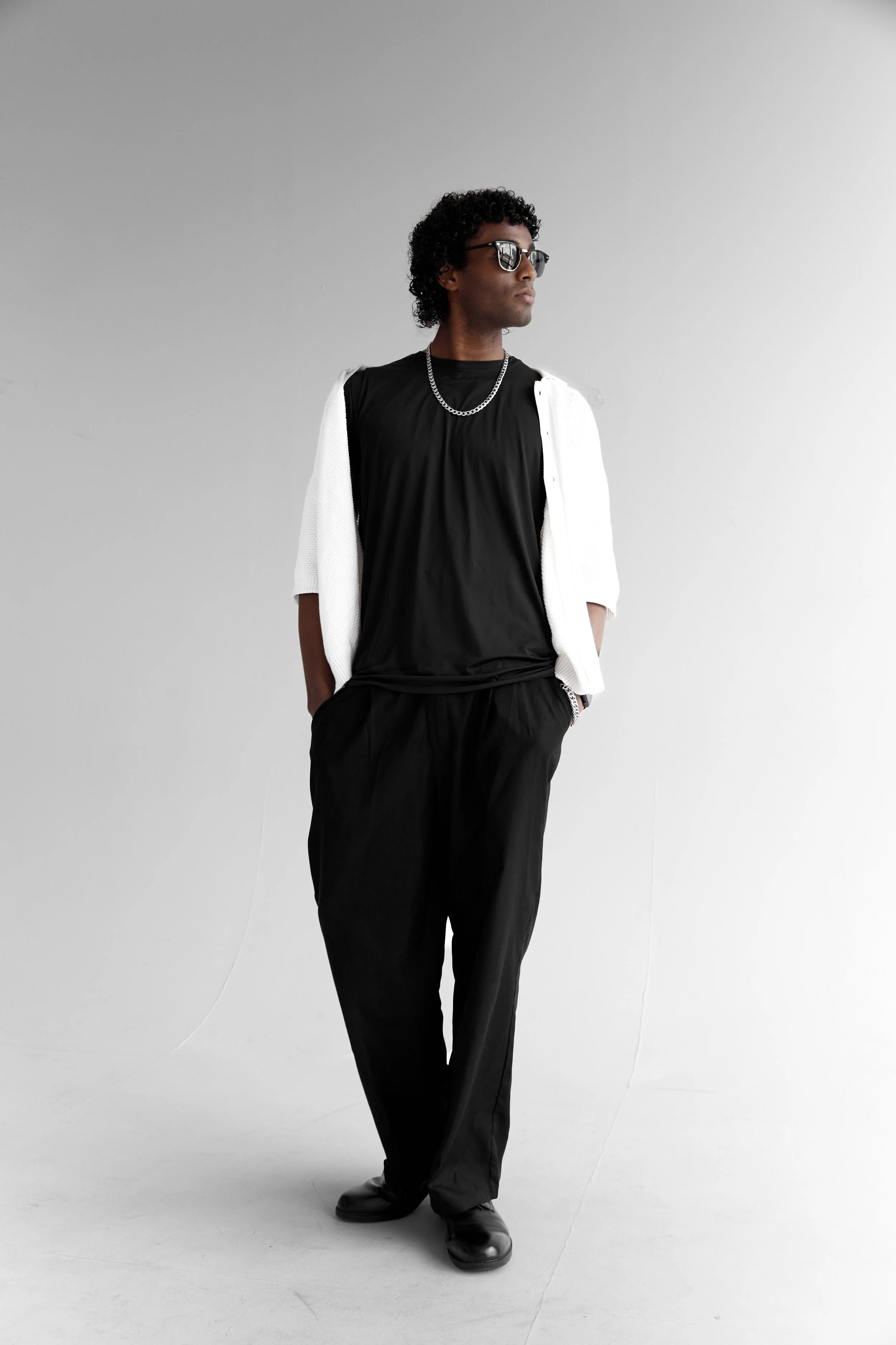 A man with curly dark hair, wearing sunglasses, a black shirt, black pants, and black shoes. He has a white jacket draped over his shoulders, a silver chain necklace, and is standing against a plain light grey background.