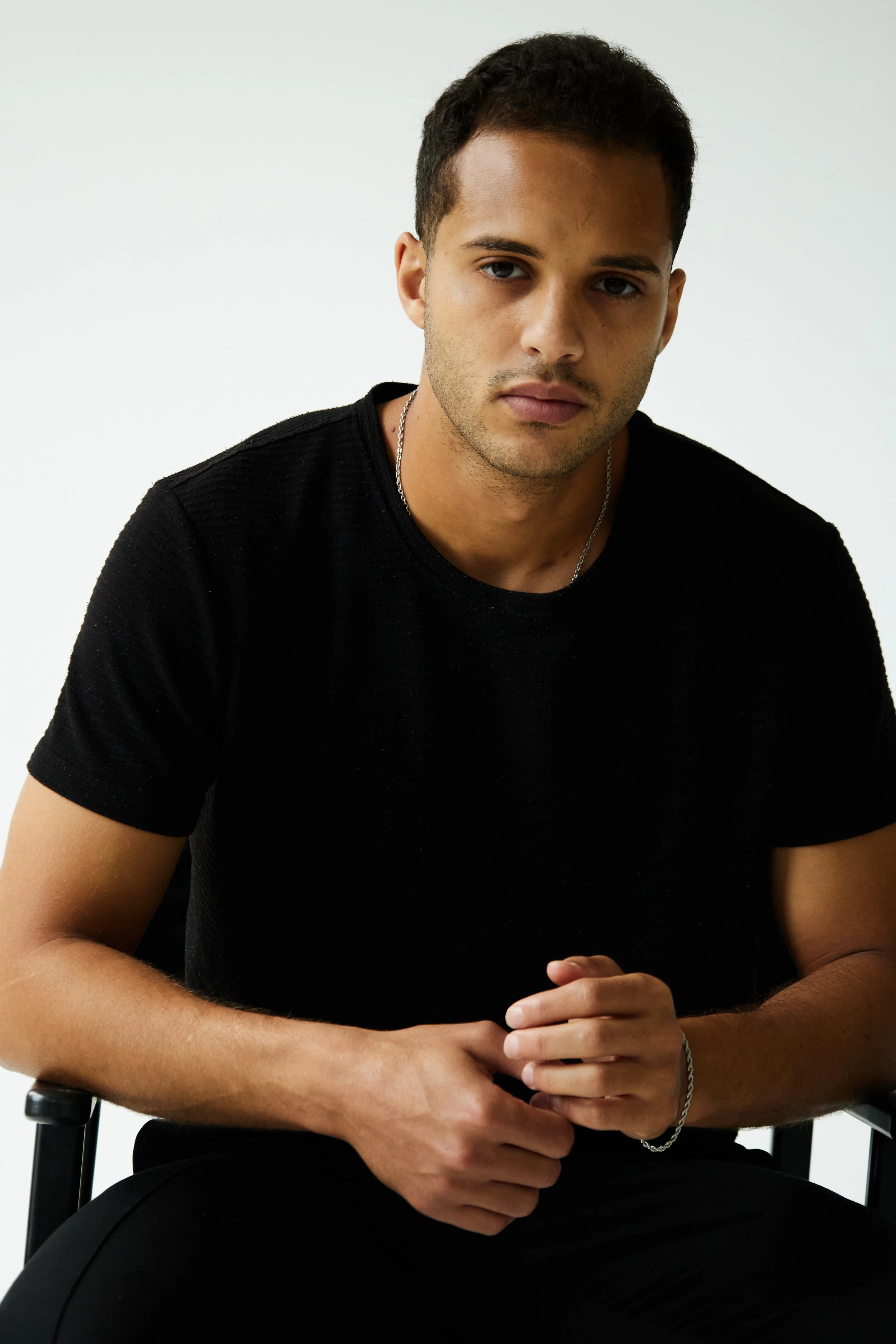 A young man sitting in a chair against a plain white background, wearing a black shirt and a silver chain, with a serious expression on his face.