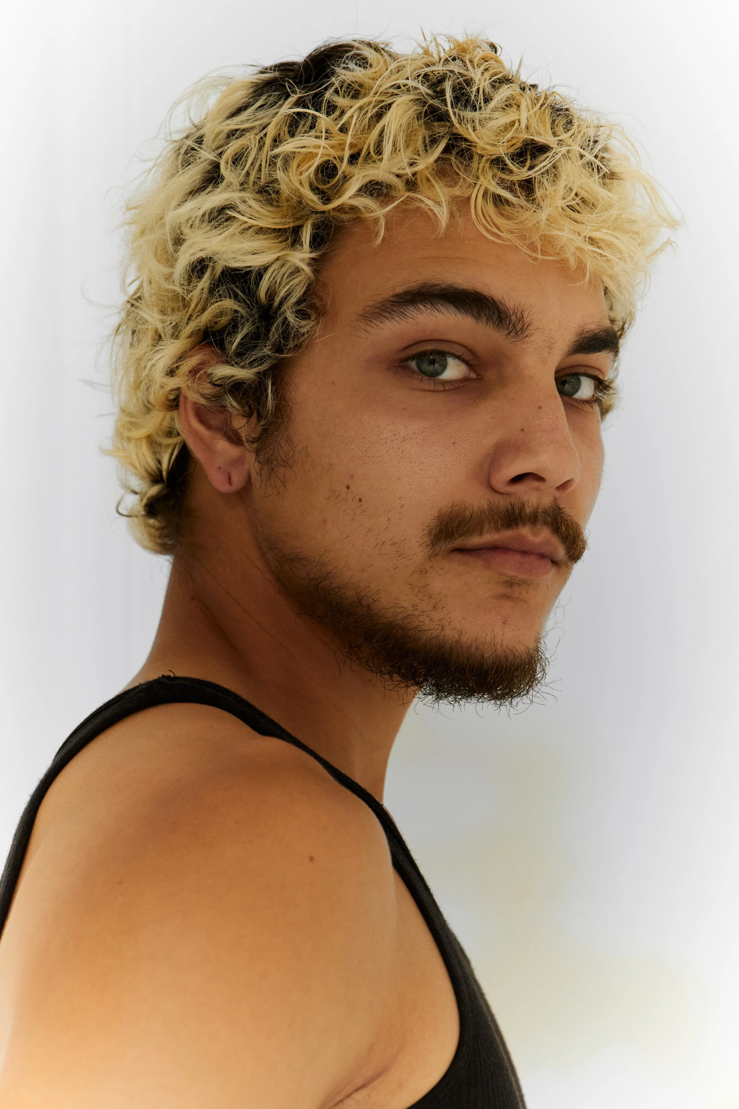 Quinton, male fashion model represented by Noirvelle Models, close-up portrait with curly blond hair against a white studio background.
