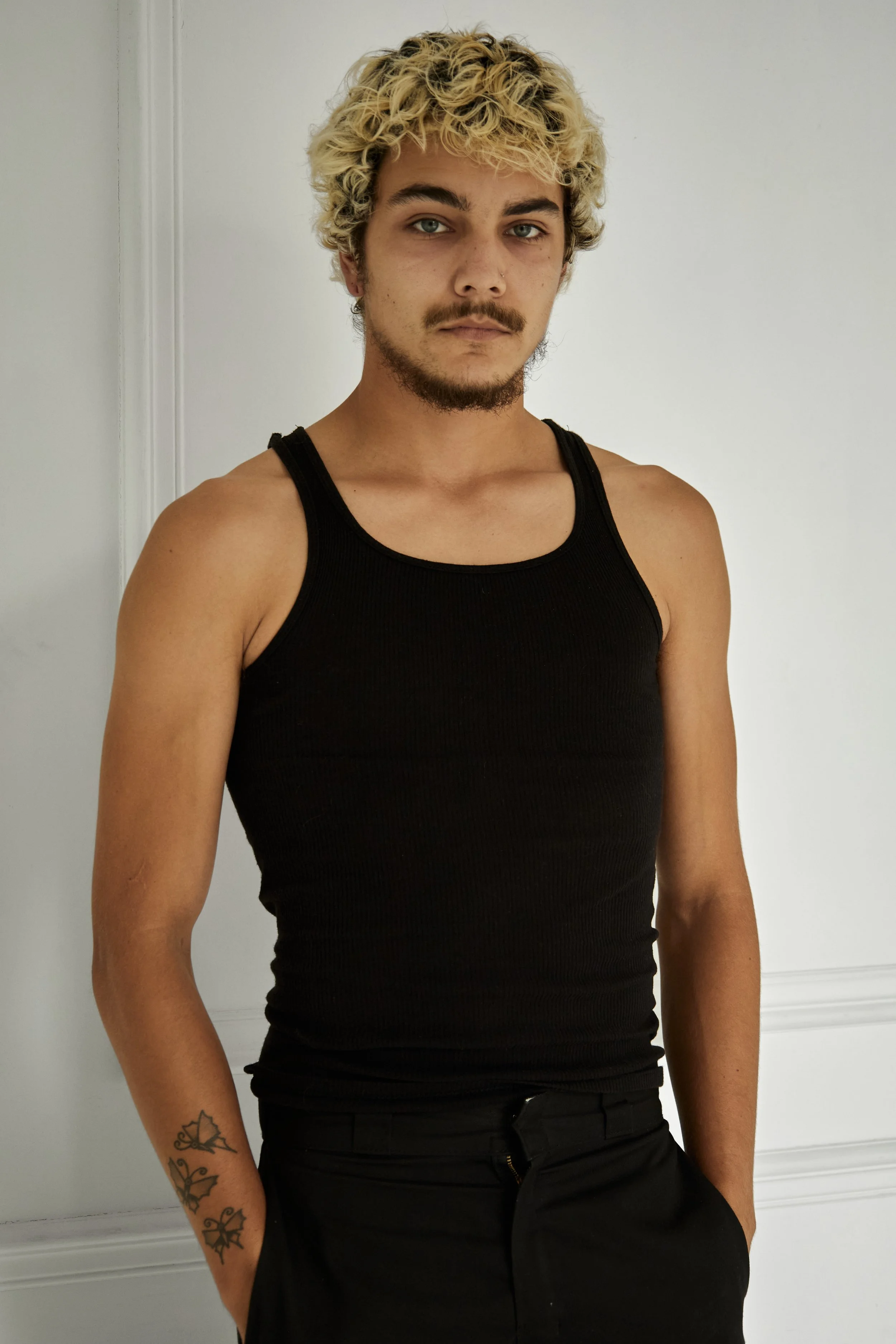 Quinton, male fashion model represented by Noirvelle Models, standing in a black tank top and black pants against a white studio background.

