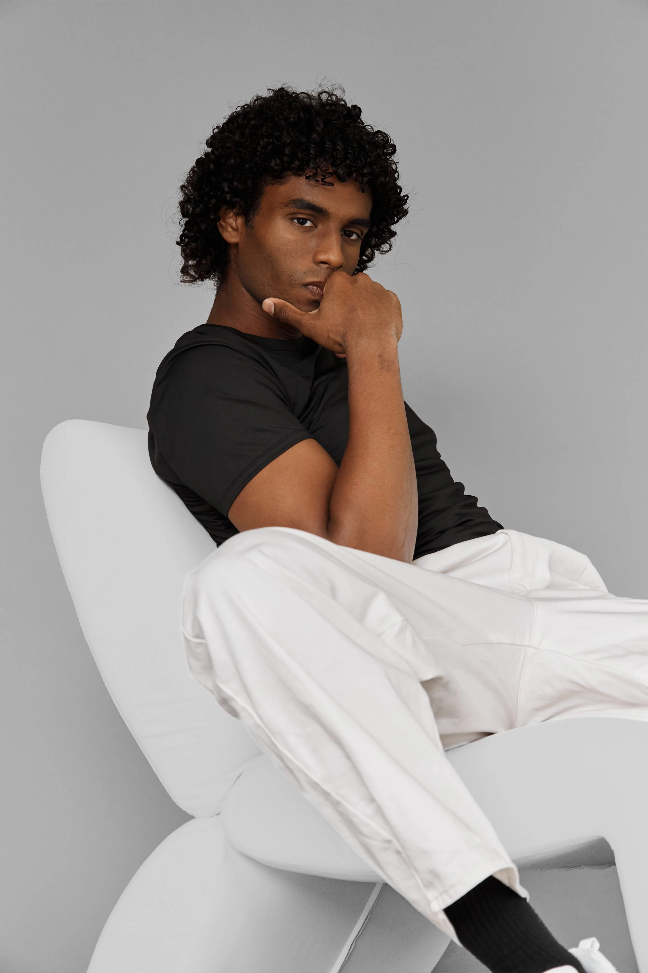  Xander, male fashion model represented by Noirvelle Models, seated in a white chair wearing a black shirt and white pants against a grey studio background.