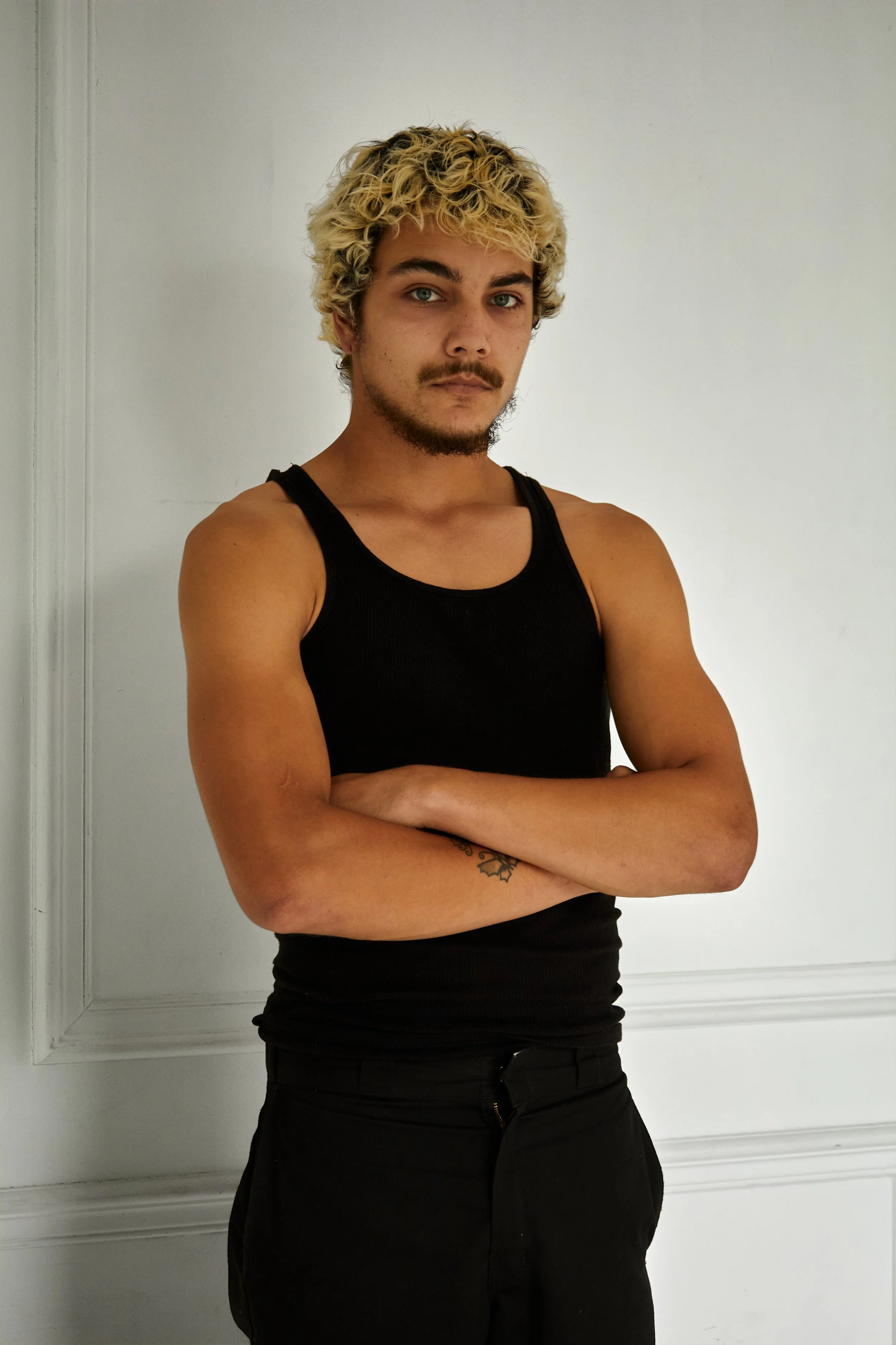 Quinton, male fashion model represented by Noirvelle Models, standing with arms crossed in a black sleeveless top against a white studio background.