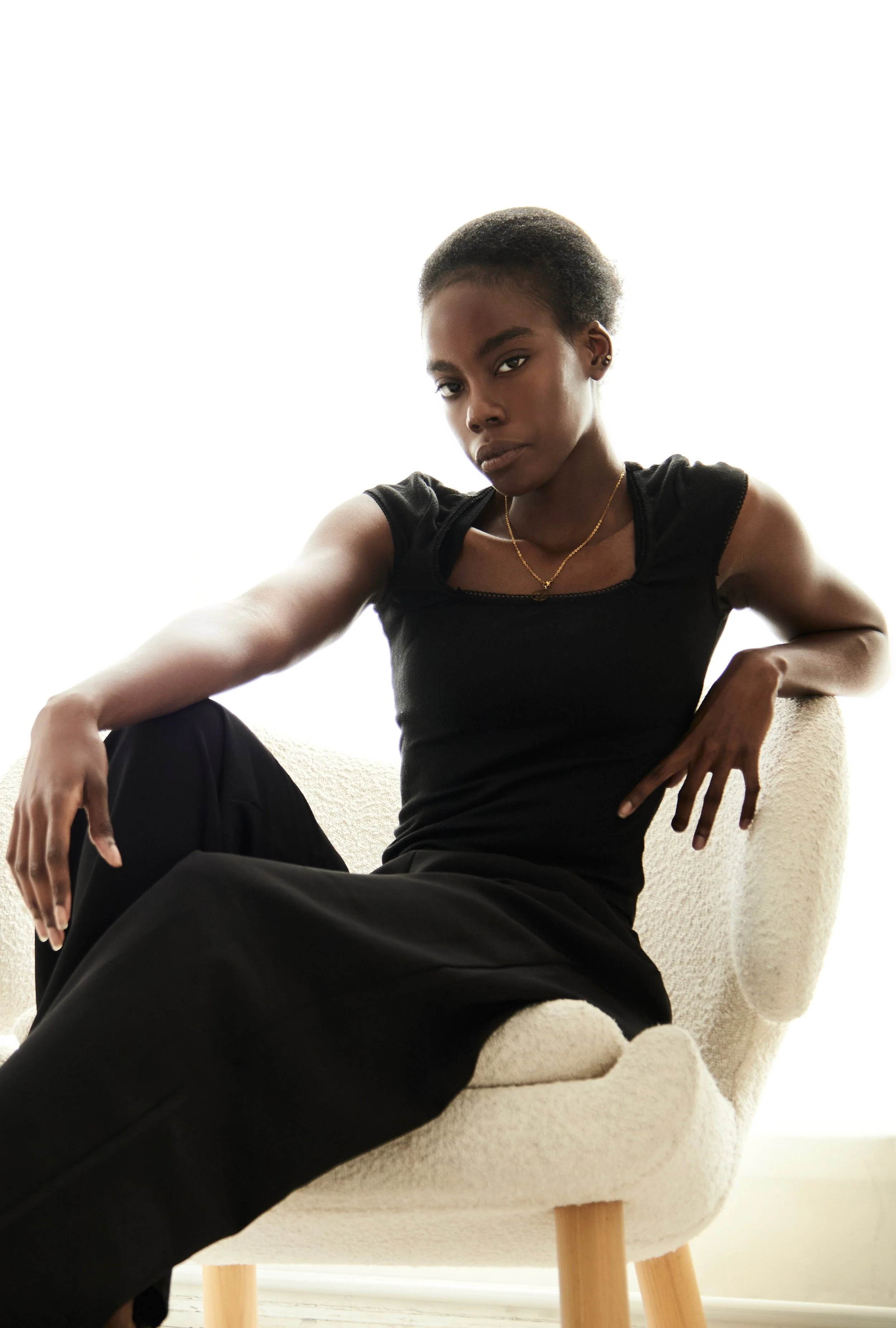 Daisy, female fashion model represented by Noirvelle Models, seated in a studio wearing a black sleeveless top and black pants.
