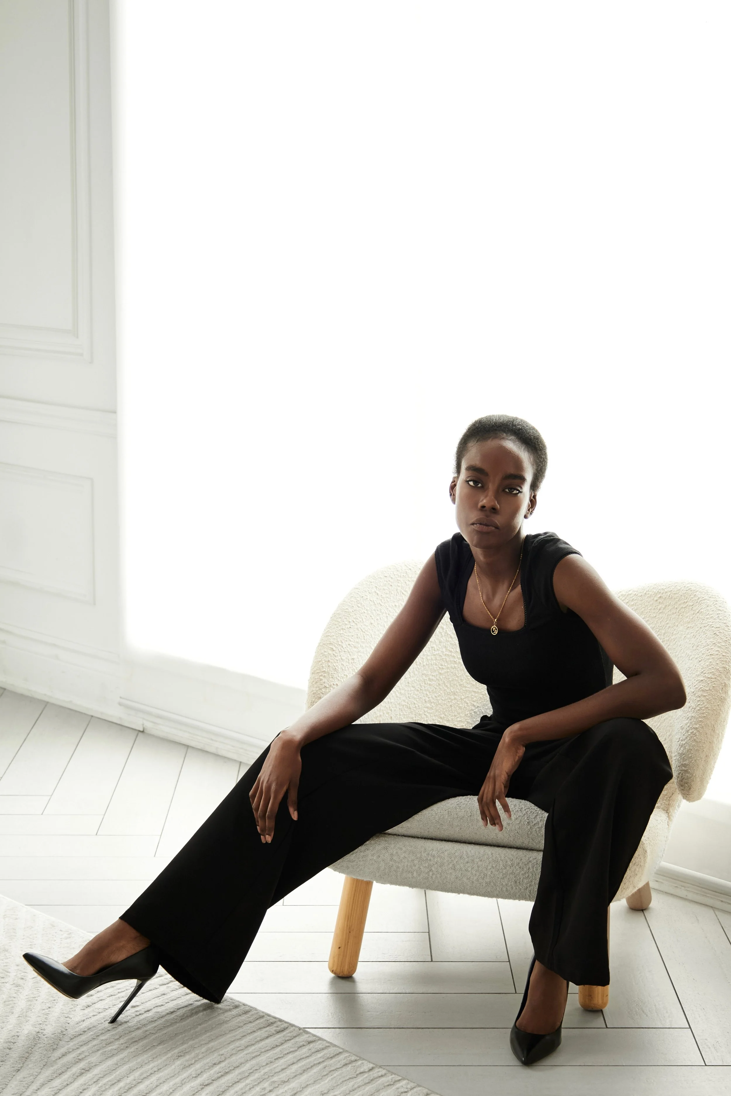 Daisy, female fashion model represented by Noirvelle Models, seated in an armchair wearing a black outfit and heels.