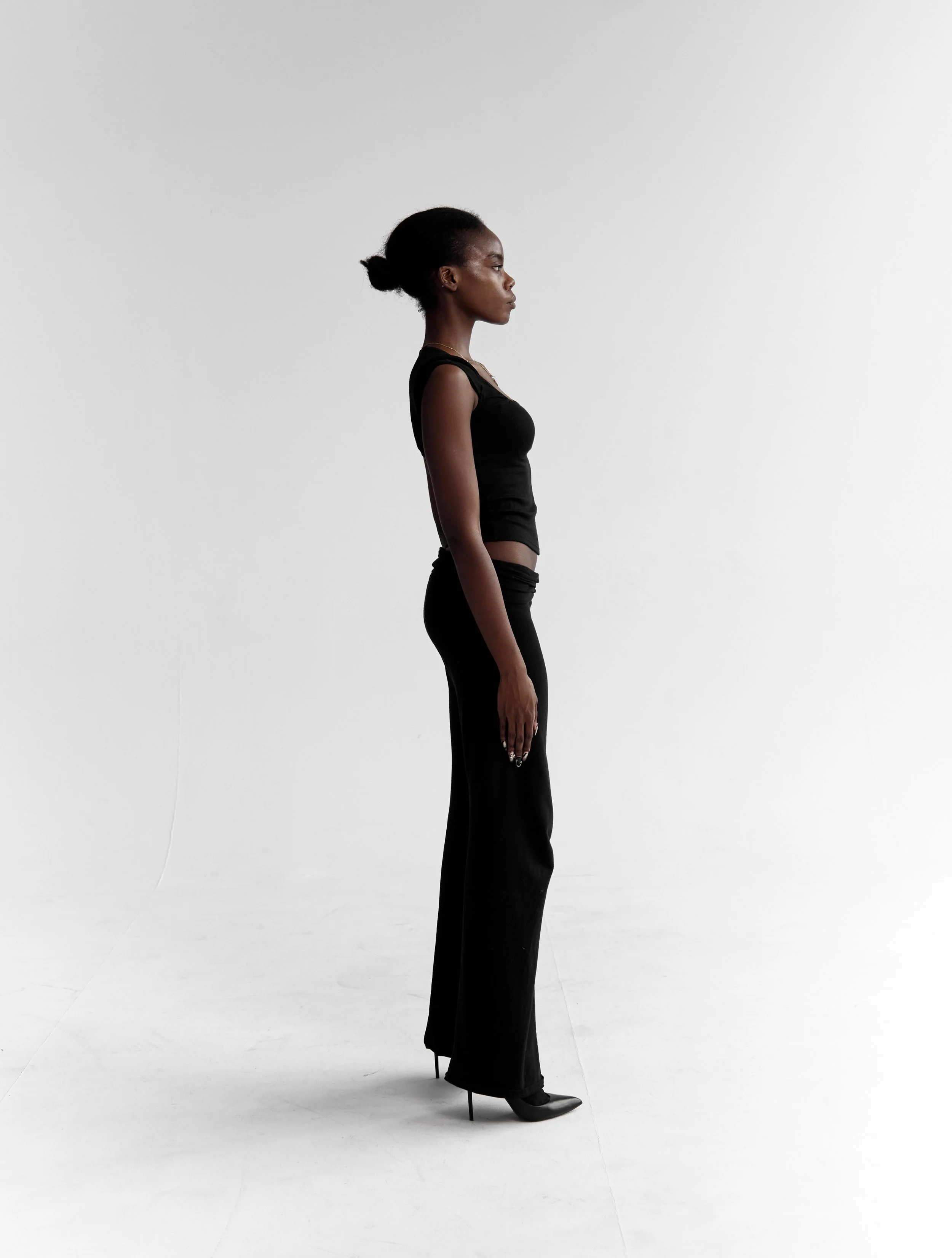  Daisy, female fashion model represented by Noirvelle Models, standing in profile in an all-black look against a white studio background.
