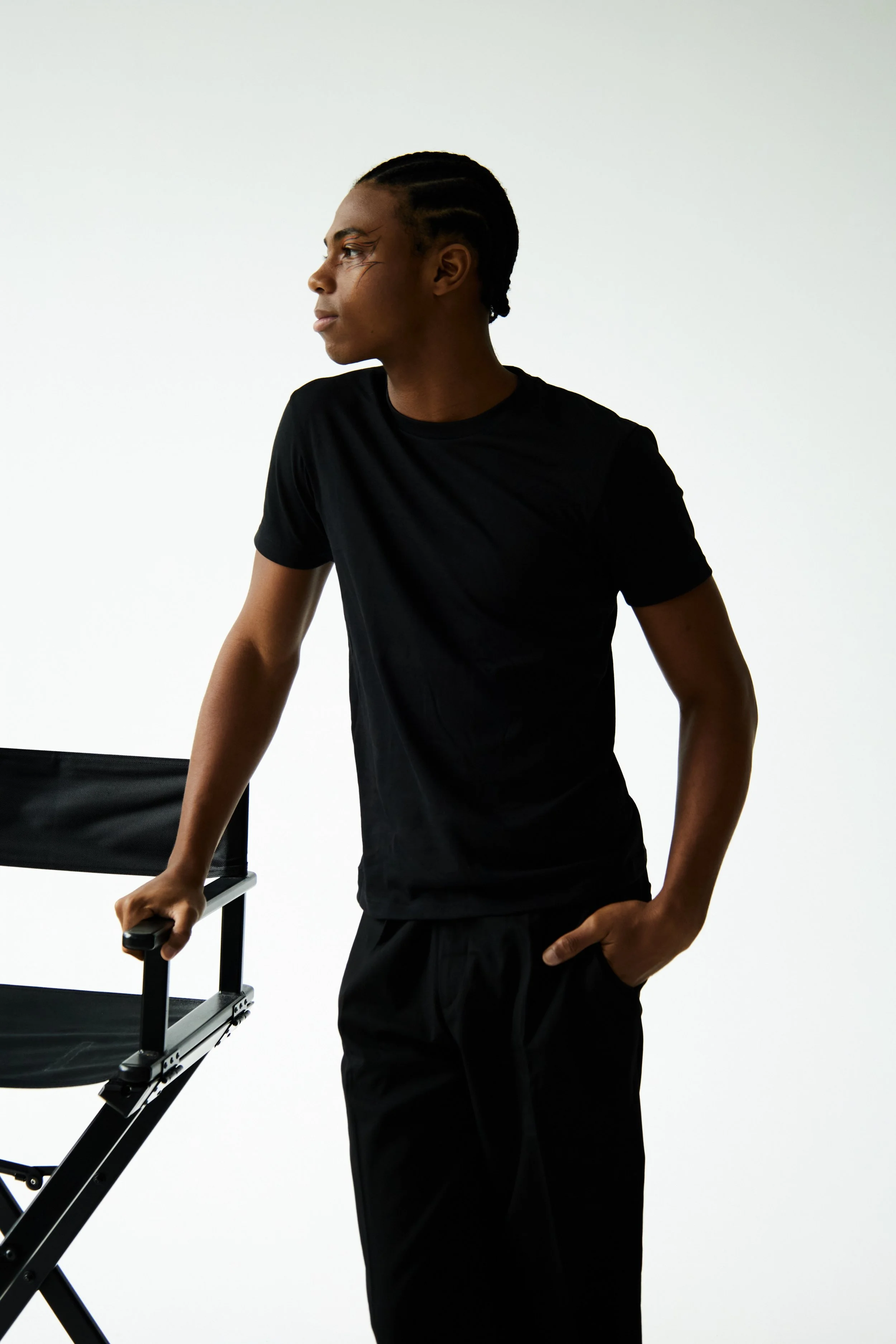 Jahvante, male fashion model represented by Noirvelle Models, posing beside a black chair in an all-black outfit against a white studio background.
