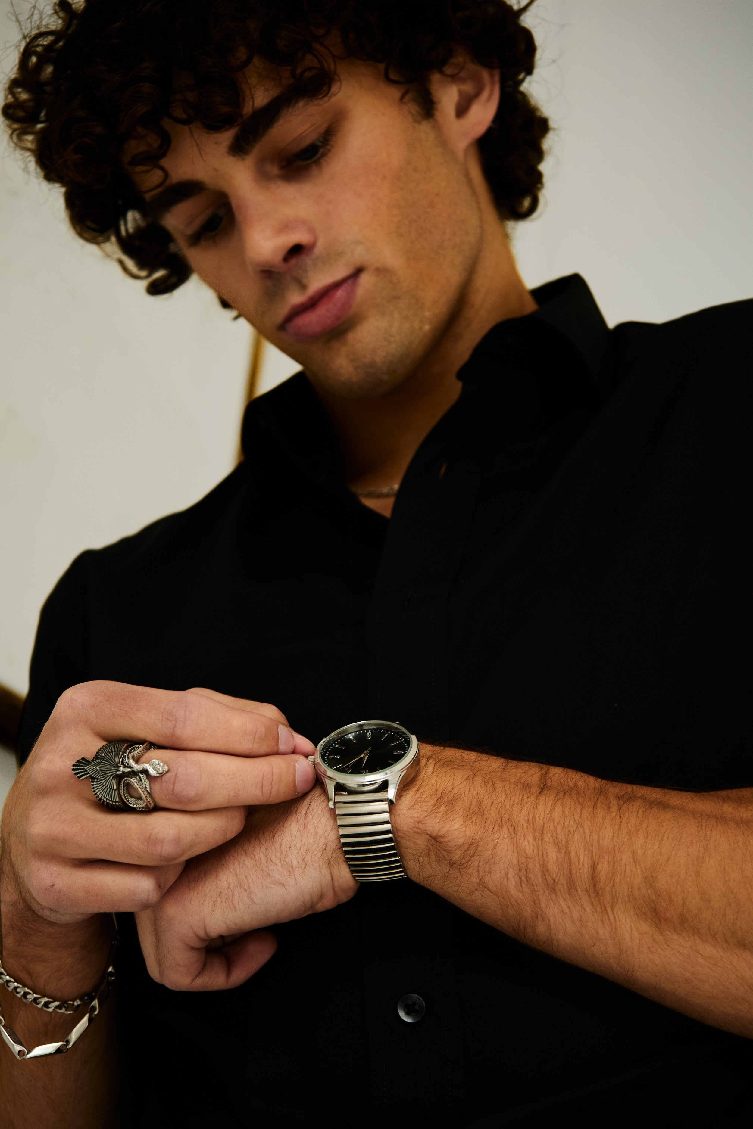 Maverick, male fashion model represented by Noirvelle Models, wearing a black look with silver jewelry and watch in a close-up fashion portrait.
