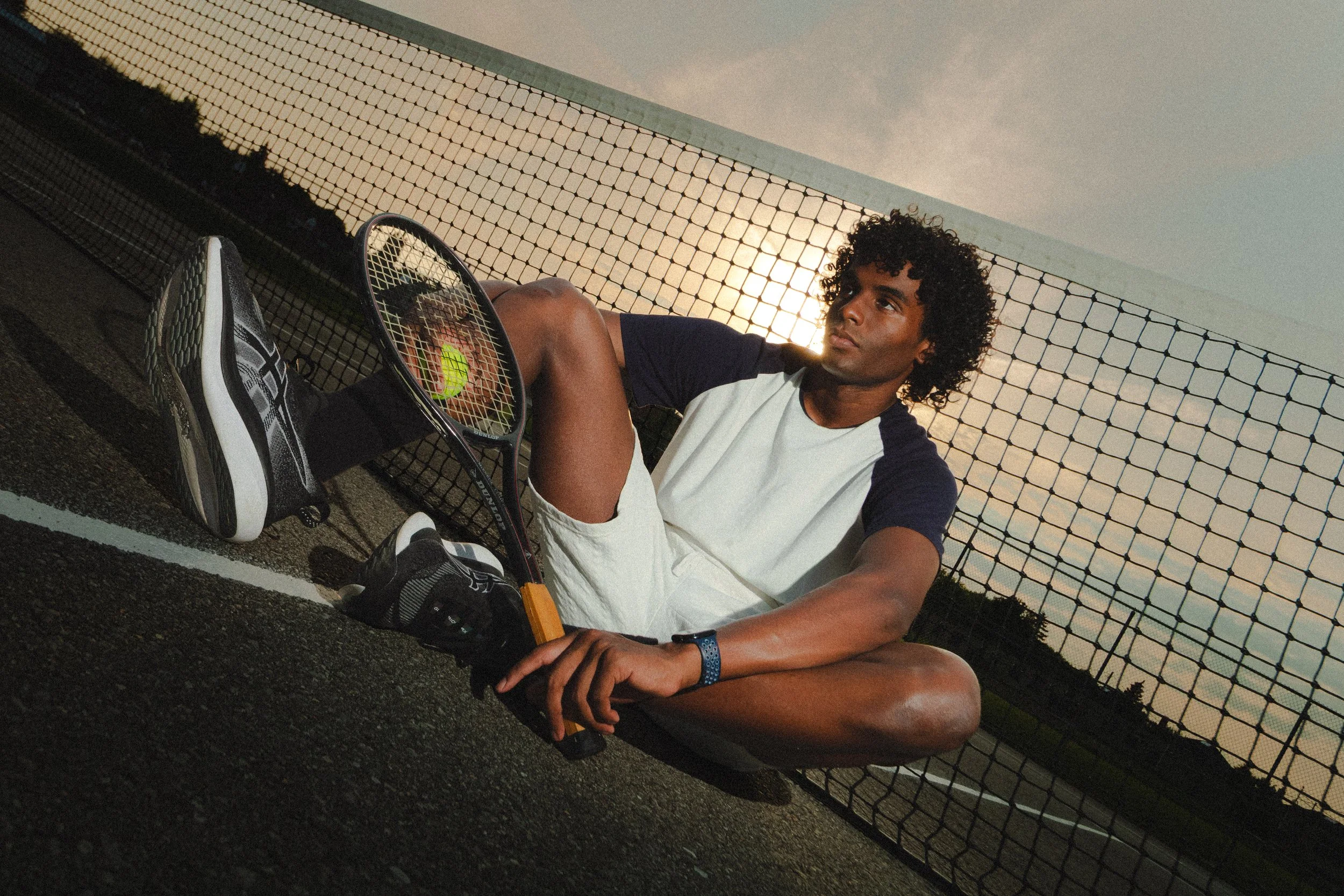 Xander, male fashion model represented by Noirvelle Models, posing with a tennis racket on a tennis court at sunset.