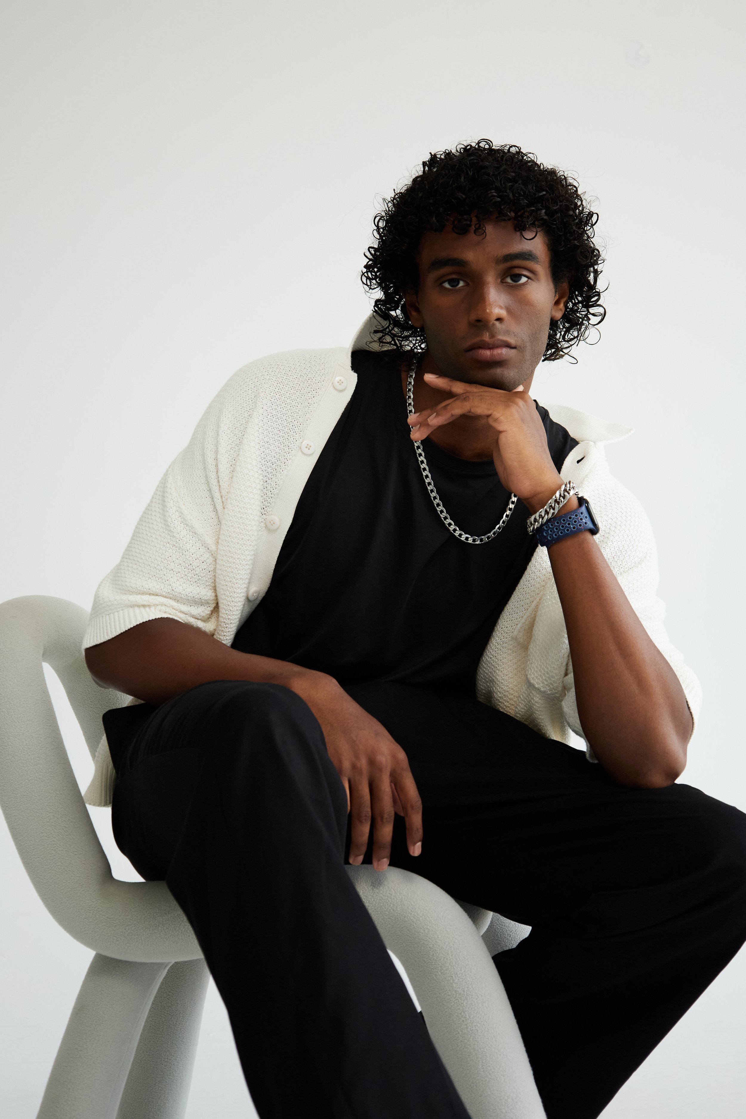 Xander, male fashion model represented by Noirvelle Models, seated in a white chair wearing a black and cream layered look.