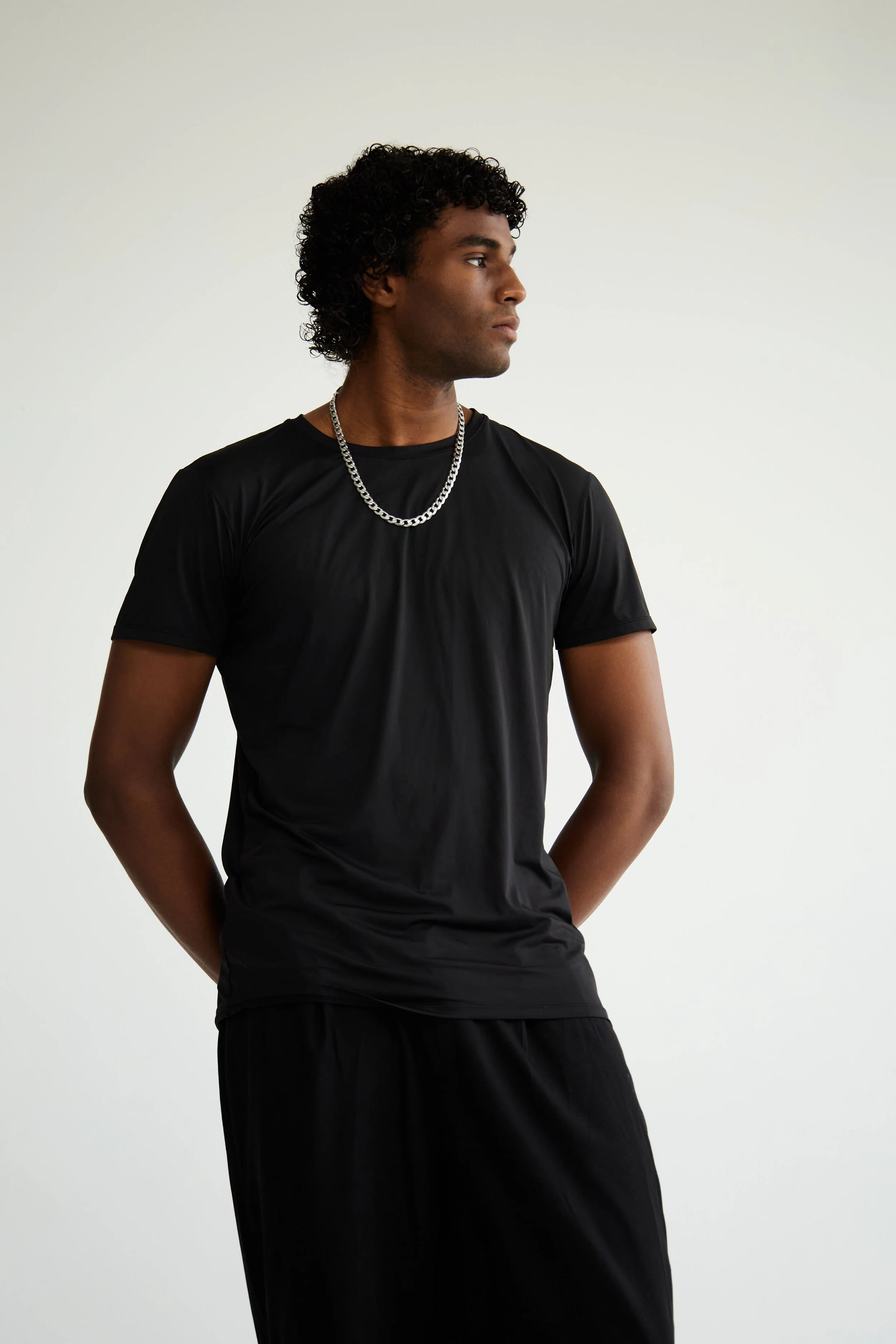 Xander, male fashion model represented by Noirvelle Models, wearing an all-black look with silver jewelry against a light studio background.
