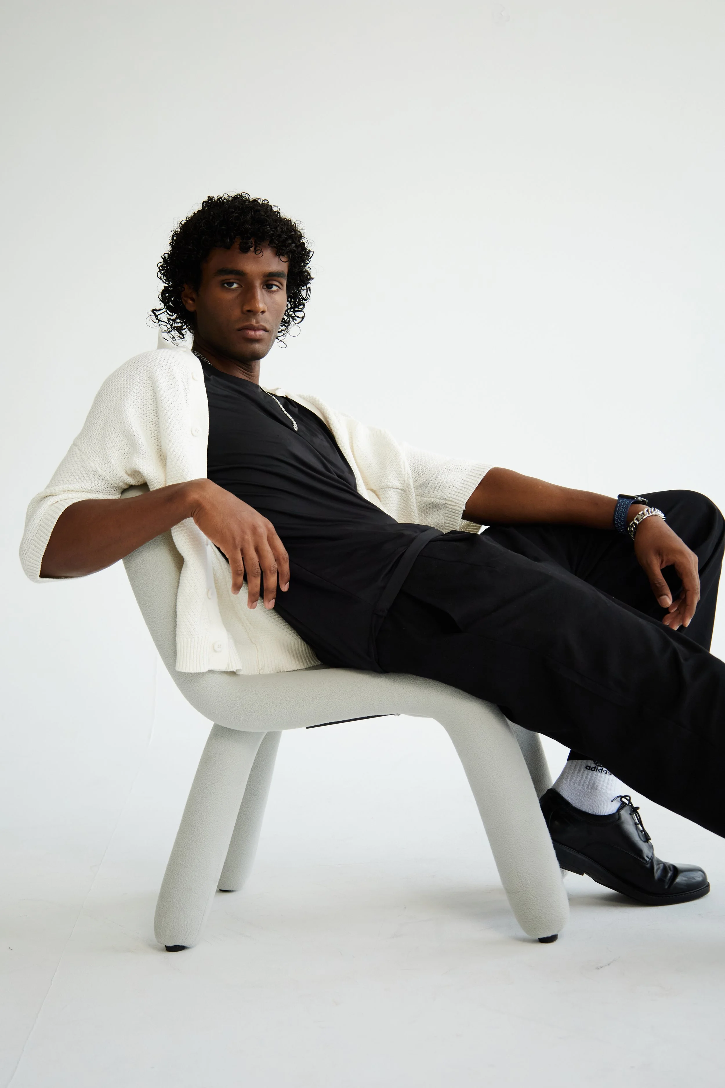 Xander, male fashion model represented by Noirvelle Models, reclining in a white chair wearing a layered black and cream look.
