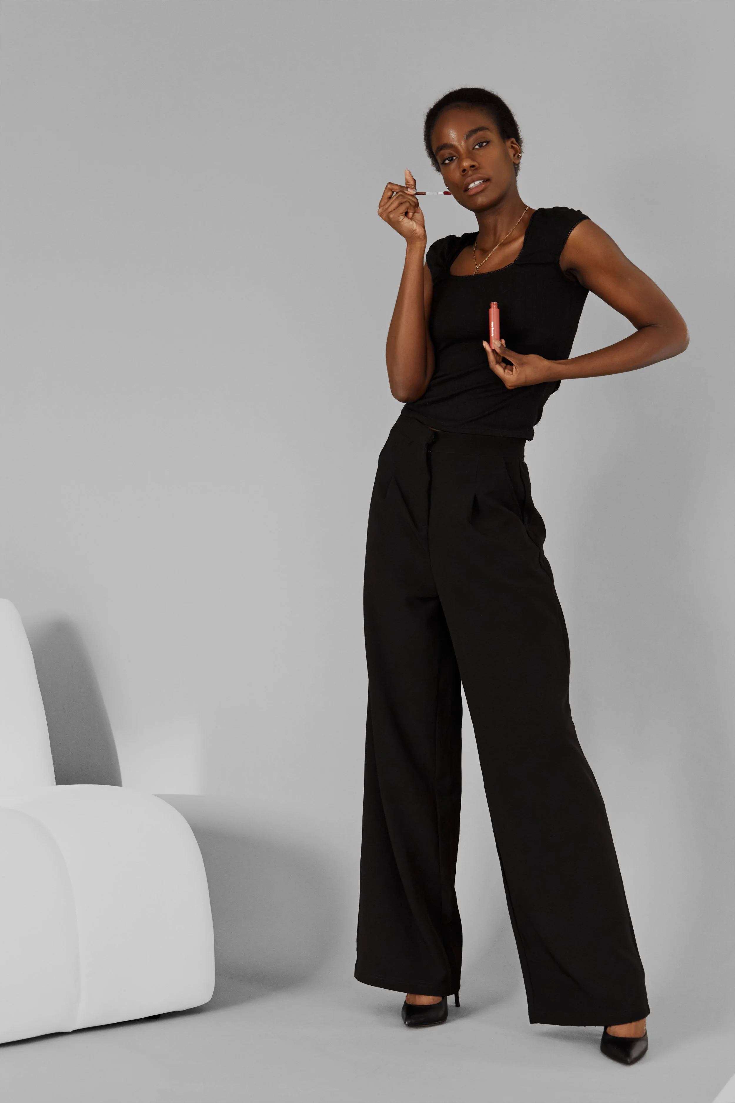 Daisy, female fashion model represented by Noirvelle Models, posing with beauty products in an all-black look against a grey studio background.
