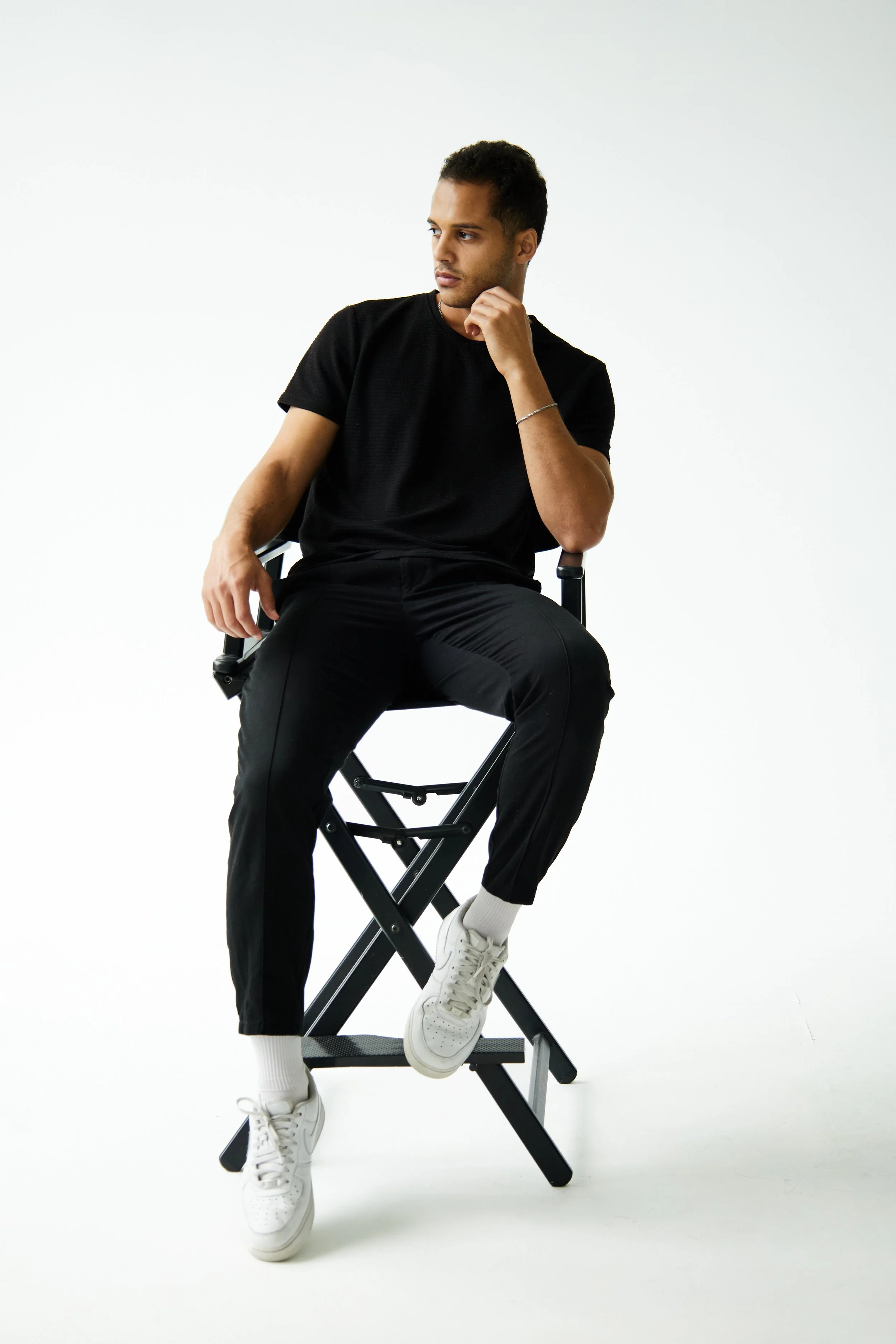 Bashir, male model represented by Noirvelle Models, posing on a director’s chair in black clothing against a white studio background.
