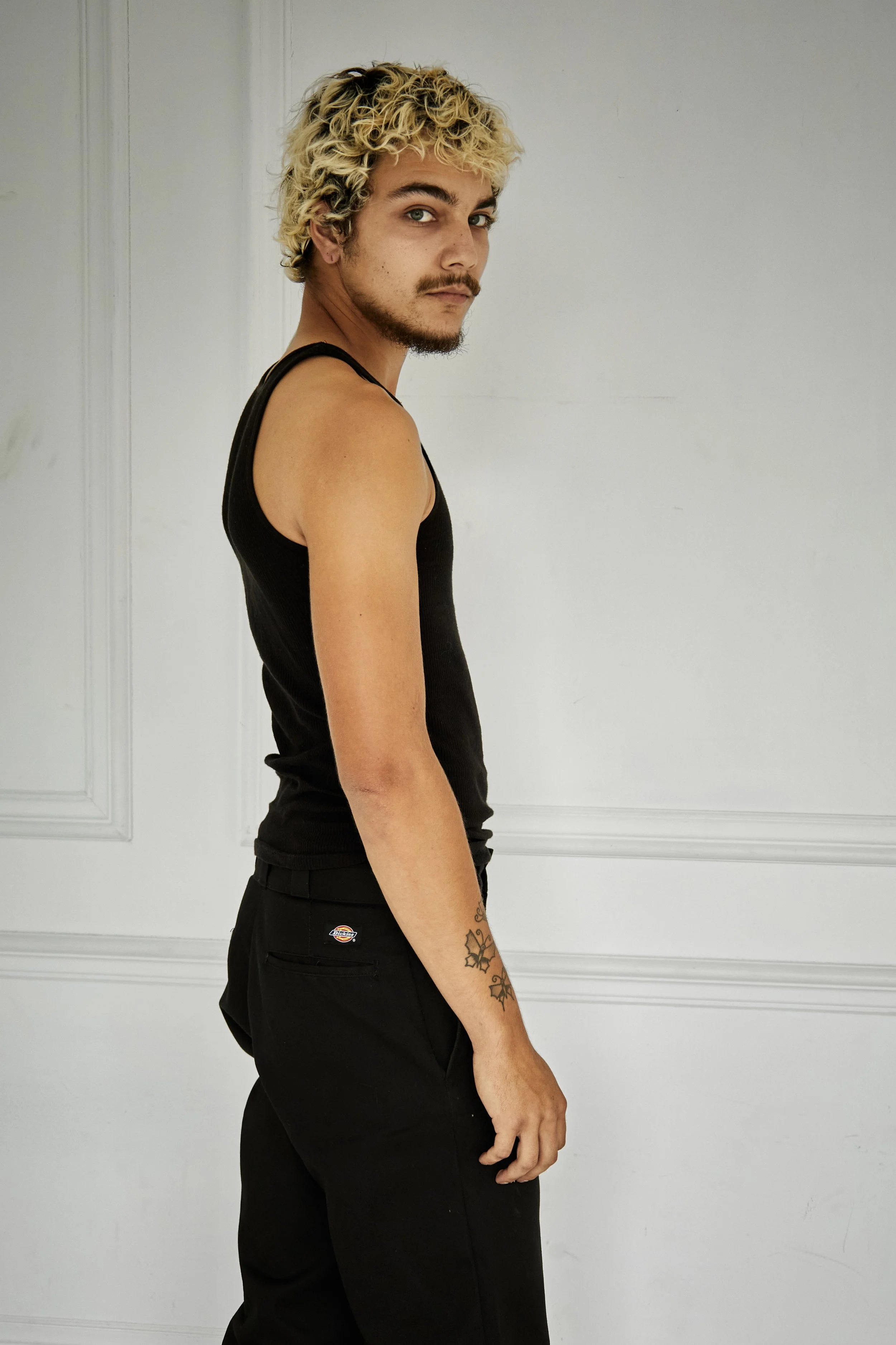 Quinton, male fashion model represented by Noirvelle Models, standing in side profile in a black tank top and black pants against a white studio background.
