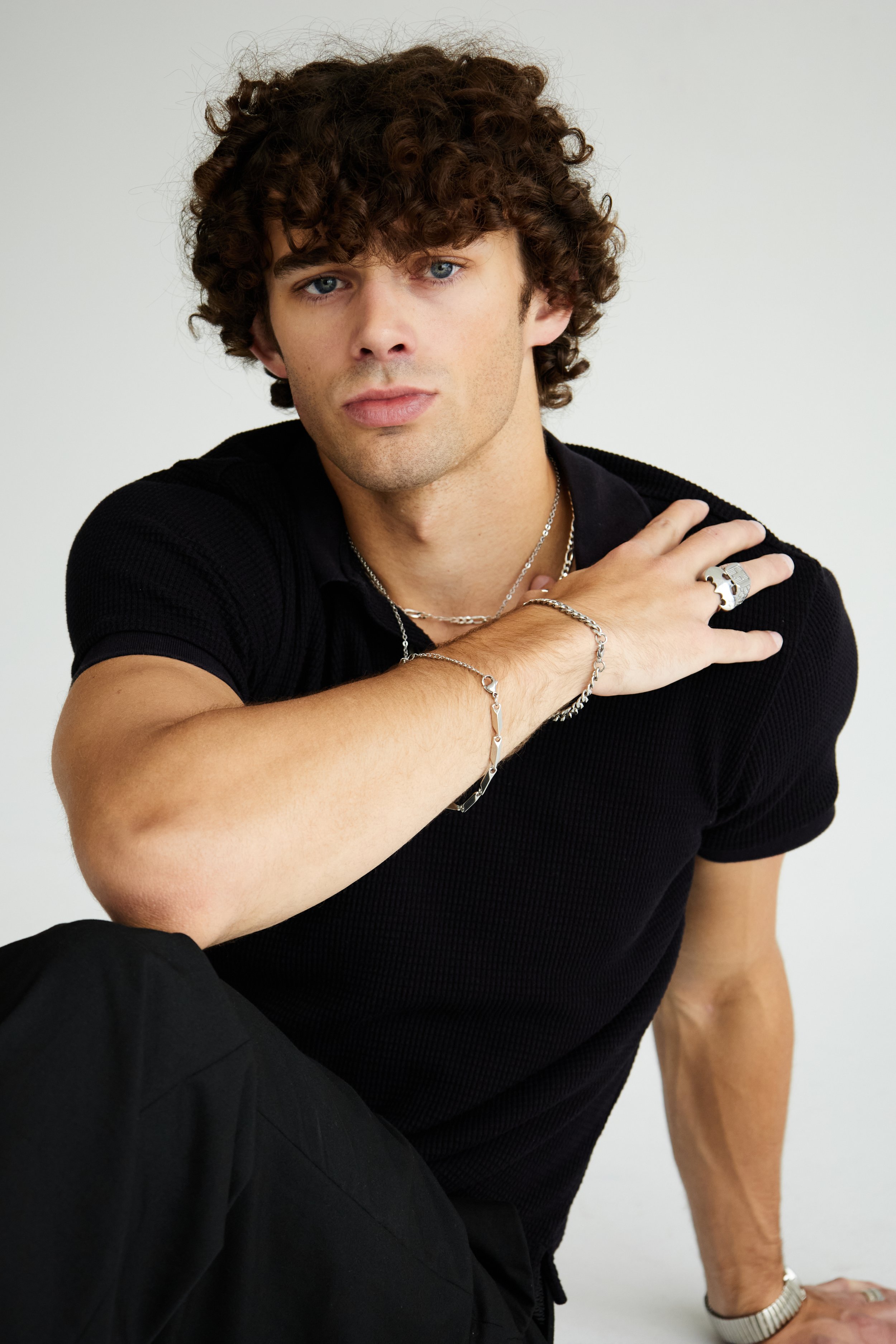 Maverick, male fashion model represented by Noirvelle Models, posing in an all-black look with silver jewelry against a white studio background.
